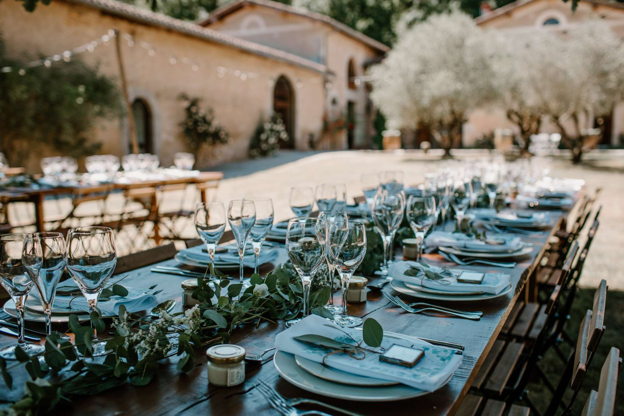 Outdoor courtyard reception table with eucalyptus runner, white plates, blue-grey napkins, and string lights