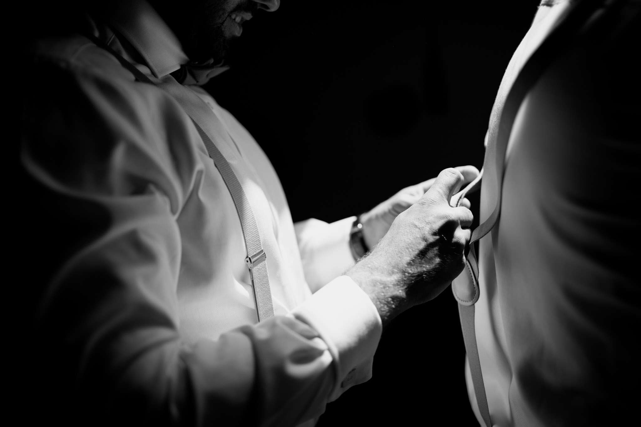 Black-and-white shot of groom being helped with suit jacket, white shirt and suspenders visible