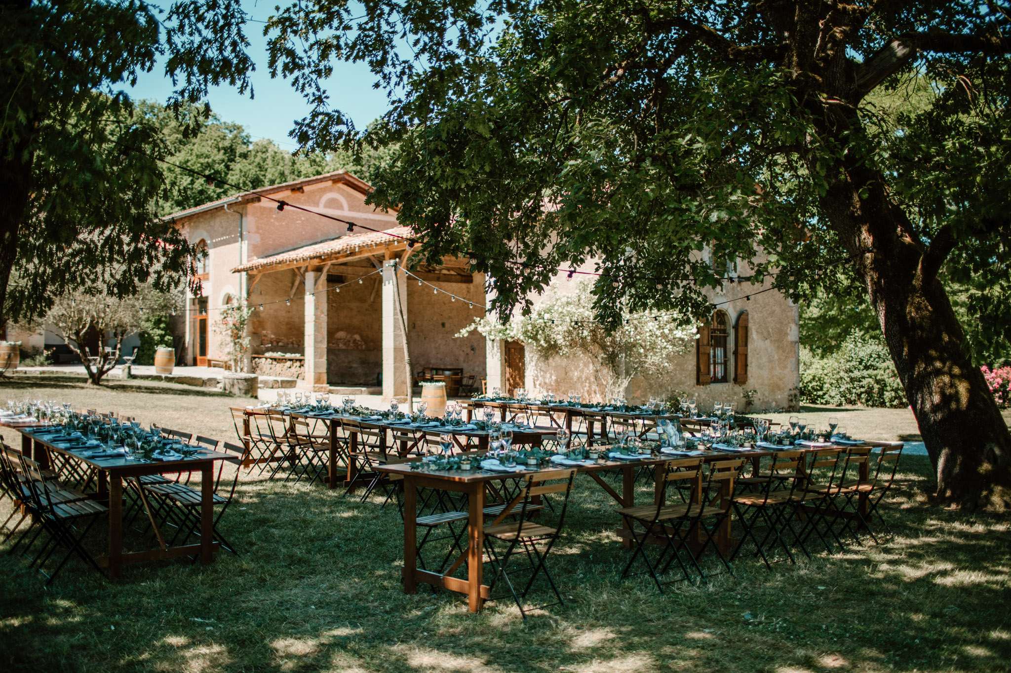 An outdoor wedding reception setup on a lawn in front of a rustic French stone farmhouse with a terracotta-tiled roof, covered terrace with columns, and wooden shutters. Four long rectangular wooden farm tables are arranged in a U-shape or parallel rows, surrounded by black folding bistro chairs. The tables are set with dusty blue-teal linen napkins, glassware, and low greenery garland centerpieces running along the center. String globe lights are strung between the trees and the building's structure, suggesting an evening ambiance is planned. A wooden wine barrel is visible near the building's terrace. Wide shot taken from under a large mature tree, capturing both the table arrangement and the venue facade in full. Potential venue feature image.