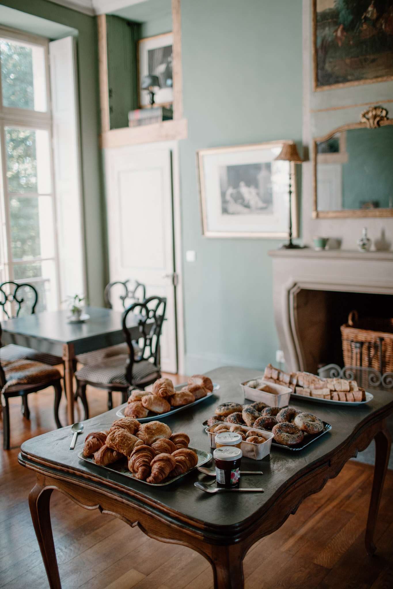 Continental breakfast on antique table in sage-walled chateau salon with marble fireplace and oil paintings