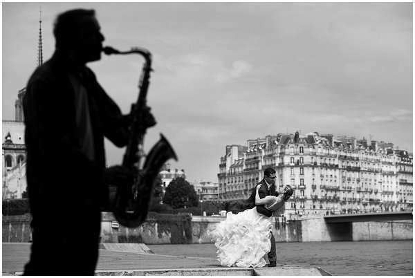 A black-and-white outdoor couple portrait taken along the Seine river in Paris, with Haussmann-style buildings and a bridge visible in the background. In the midground, the groom dips the bride dramatically, her full ruffled ballgown skirt fanning out toward the ground. In the foreground, a saxophonist is captured in sharp silhouette, playing while facing left, creating a strong compositional layering effect. The image is a wide environmental shot with high contrast between the dark silhouetted figure and the lighter tones of the Parisian skyline behind the couple.