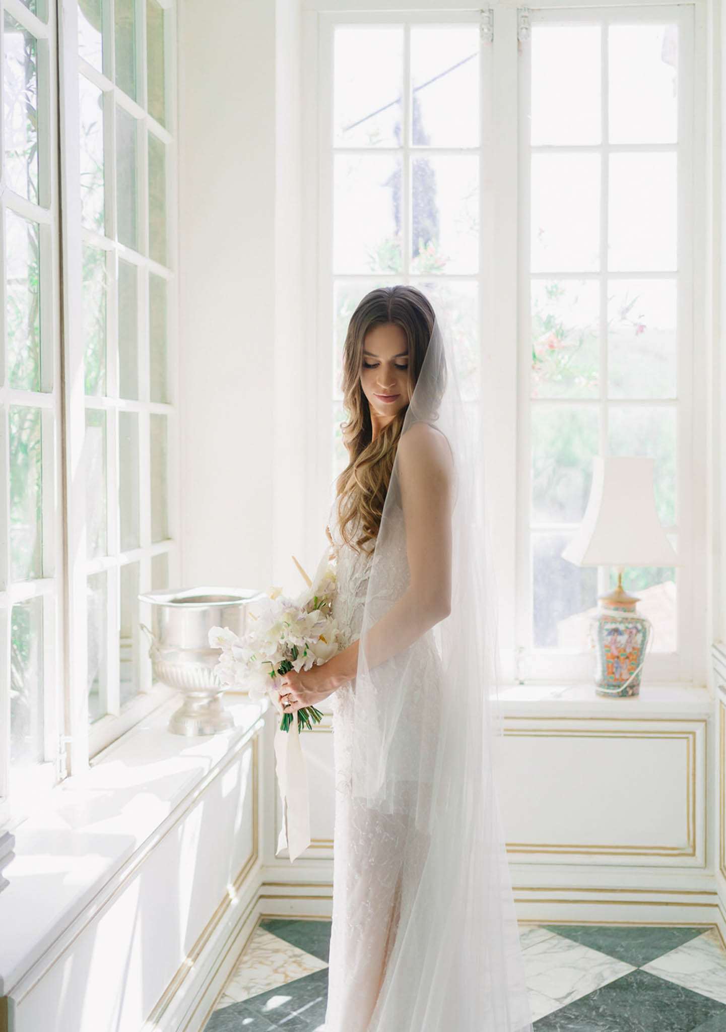 A bridal portrait taken indoors in a bright, ornate room with white and gold paneled walls, tall French windows, and a green and white diamond-patterned marble floor. The bride stands alone, looking downward, wearing a fitted ivory lace gown with a long cathedral-length tulle veil draped over one shoulder. She holds a compact bouquet of white orchids and ivory blooms with green foliage. Her dark hair falls in loose waves past her shoulders. The room is decorated in a classic French interior style, with a decorative porcelain lamp on a white and gold console table and a silver urn-style vessel on the windowsill. The shot is a full-length portrait, backlit by natural light flooding through the windows.