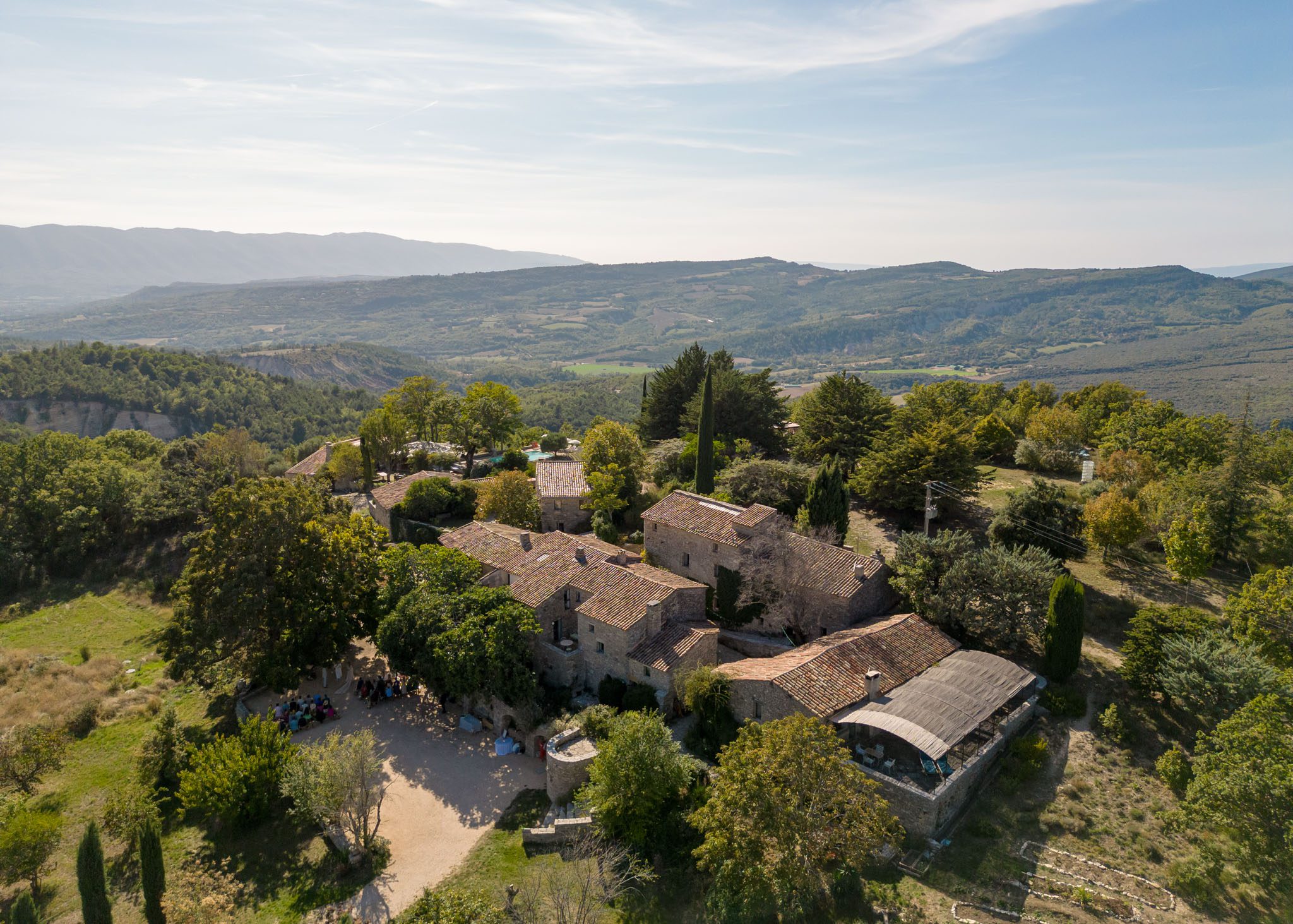 Aerial view of Provencal stone mas with terracotta roofs on hillside overlooking forested valley with guests in courtyard