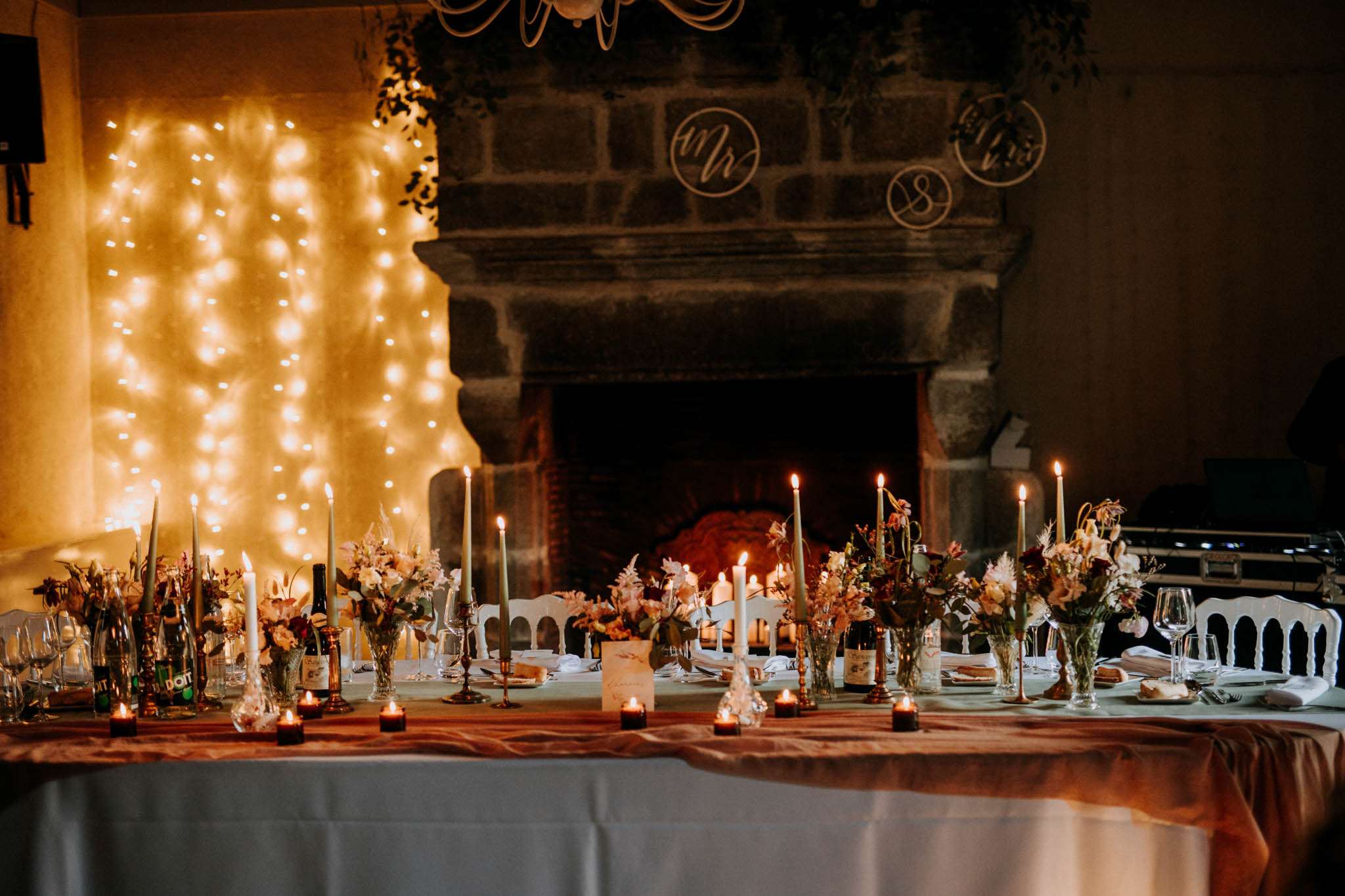 A wide shot of a decorated wedding reception dinner table set inside a room with a large stone fireplace as the focal backdrop. The table is dressed with a white linen tablecloth and a terracotta-rust colored satin runner, with multiple clusters of floral arrangements featuring blush, rust, and warm peach tones mixed with greenery, placed in glass vases. Tall sage green taper candles in brass candlestick holders are distributed along the table's length, accompanied by small tea light candles in dark votives at the table's edge. Wine bottles, glassware, and white folded napkins complete the place settings, along with a visible table name card. Behind the table to the left, a curtain of warm white fairy lights creates a glowing backdrop. The stone fireplace surround features circular wire monogram signs reading 'Mr' and 'Mrs' along with what appears to be a number or initial, and the fireplace itself is lit with a visible flame. White French-style chairs are partially visible at the right. The overall decor palette is warm and earthy with terracotta, sage, and brass tones in a classic-rustic style.