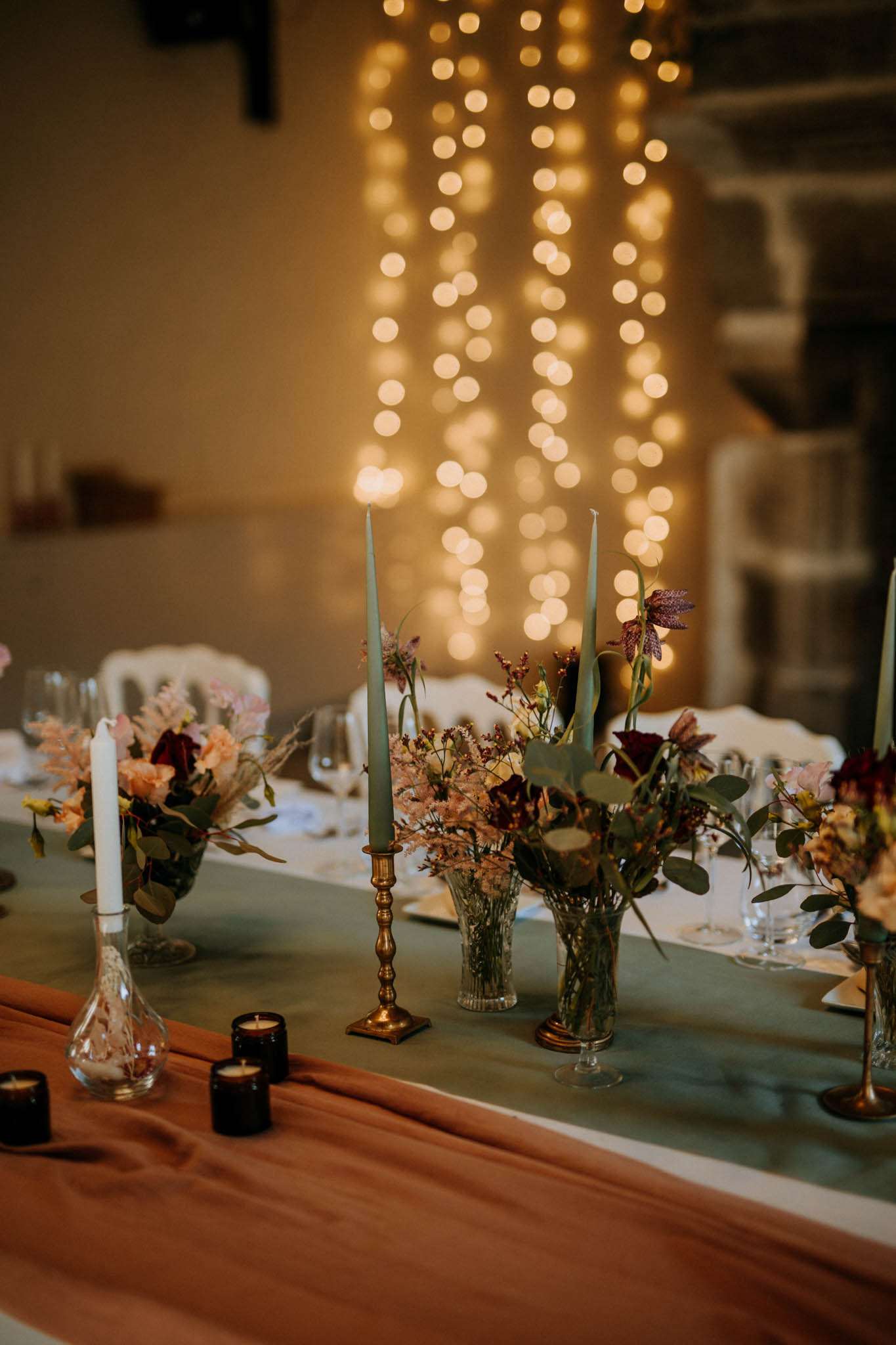A detail close-up shot of an indoor wedding reception table setup. The long table is dressed with a sage green table runner layered over a terracotta-colored fabric, with small black glass tea light holders placed along the edge. Centerpieces consist of mixed low floral arrangements in glass vases featuring dusty pink dried-style blooms, burgundy flowers, lavender sprigs, and eucalyptus foliage. Tall sage green taper candles are displayed in brass candlestick holders, alongside a single white taper in a slender glass vase. Wine glasses and place settings are visible along the table length. In the background, cascading warm-toned fairy lights create a heavily bokeh'd curtain of light against an interior wall, with white chairs partially visible. The overall styling palette combines sage green, terracotta, brass, and deep burgundy in a romantic, earthy aesthetic.