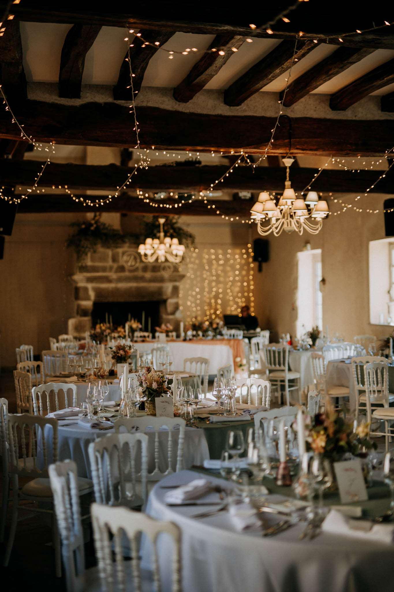 A wide interior shot of a wedding reception room set for dinner, with no guests present. The room features exposed dark wooden ceiling beams strung with multiple strands of warm fairy lights, two white chandelier-style pendant lights, and a backdrop of a dense fairy light curtain behind the head table positioned in front of a large stone fireplace. Round guest tables are dressed in white linen tablecloths and surrounded by white Napoleon/chiavari chairs, set with glassware, folded white napkins, and small floral centerpieces in muted tones of blush, rust, and dusty mauve. A long rectangular head table with a blush-toned runner is positioned against the back wall beneath the fireplace. The overall decor palette is warm white and gold with romantic rustic elements, mixing the exposed beam architecture with refined table settings. Potential venue feature image.