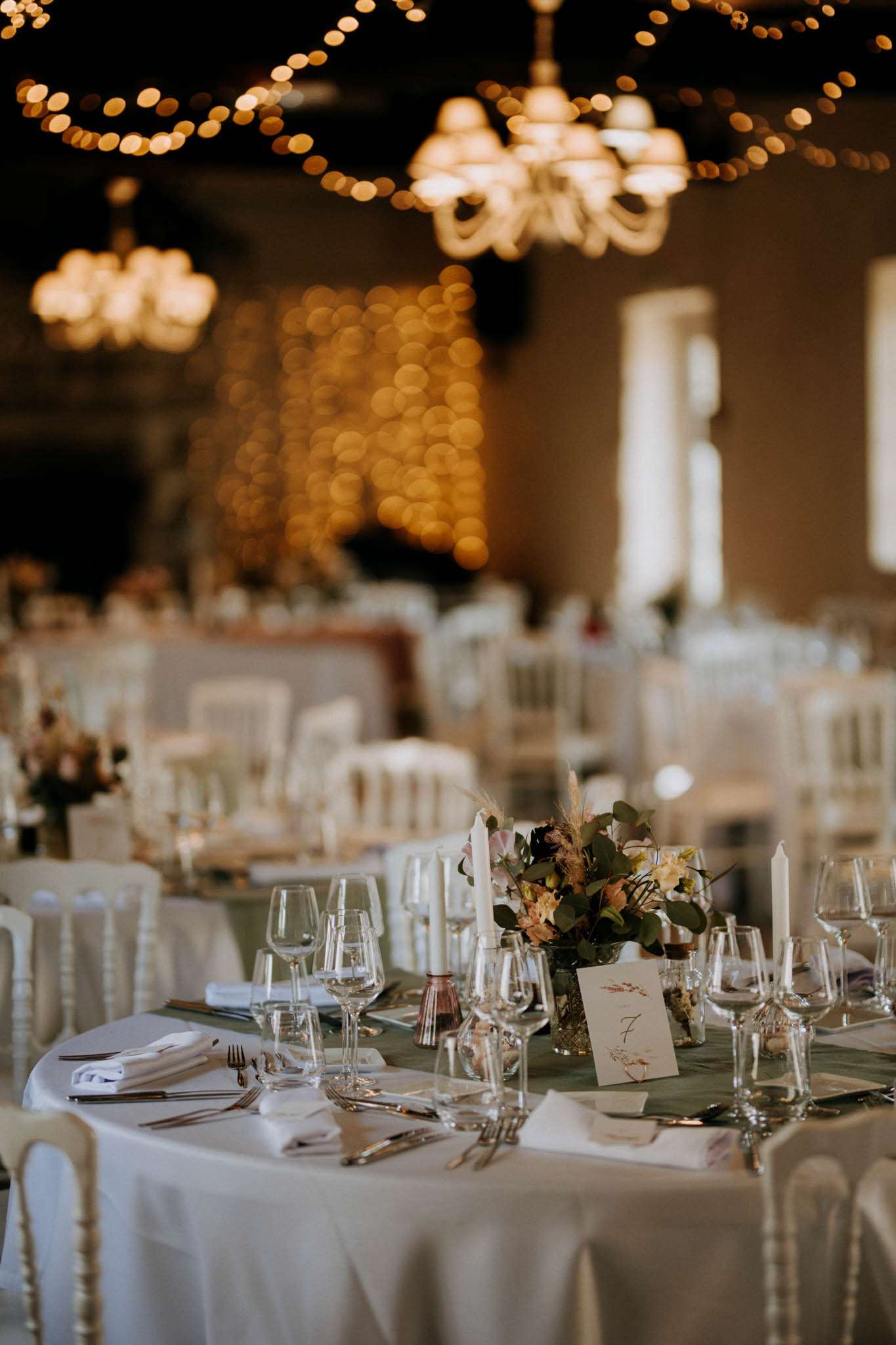 Reception table with sage green runner, peach and blush floral centerpiece, white Chiavari chairs, fairy-lit hall