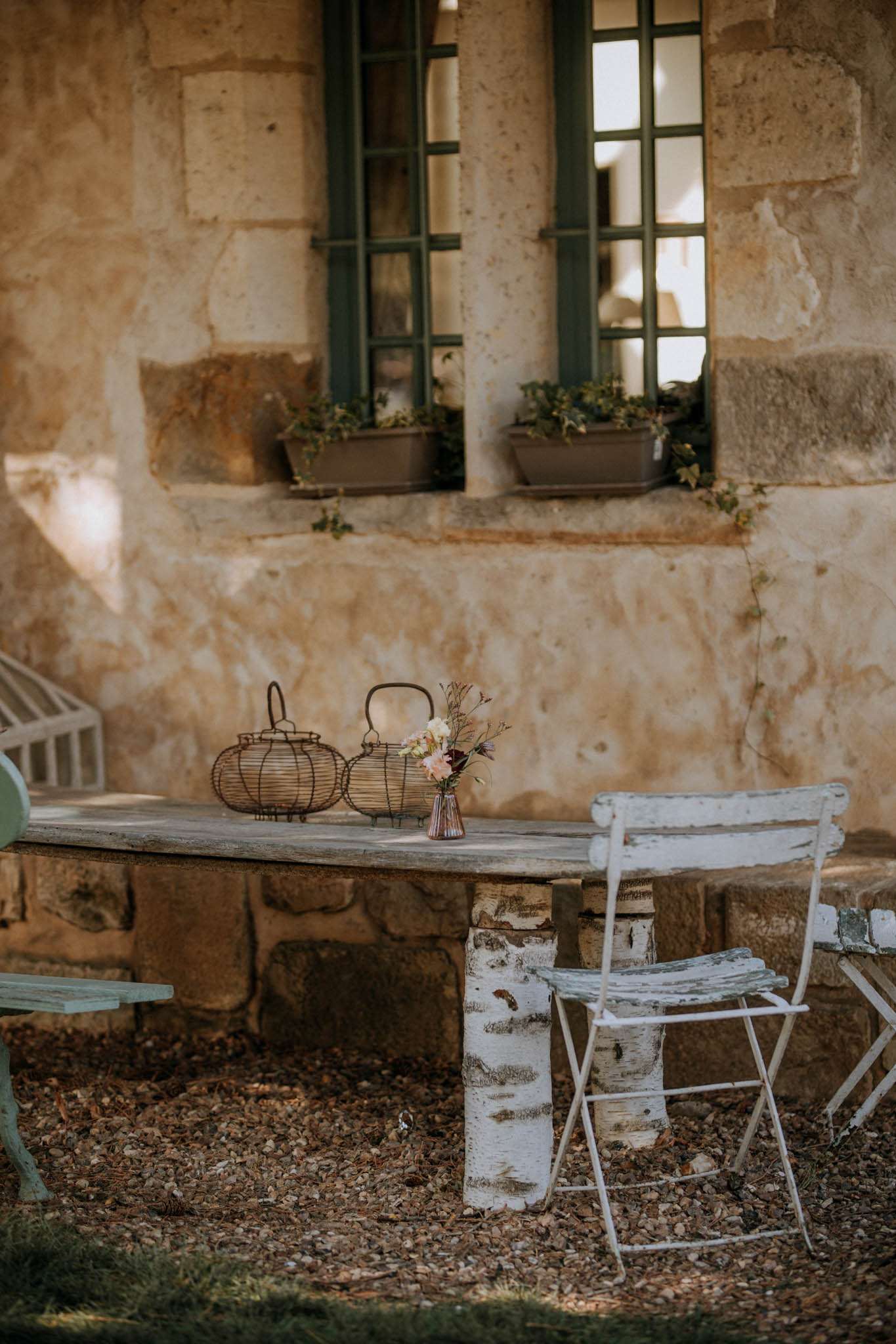 An outdoor detail shot featuring a rustic wooden table with birch log legs, styled with two wire lanterns and a small glass bud vase holding a loose arrangement of blush pink, burgundy, and mauve wildflowers. The table is flanked by distressed white metal folding chairs and a pale mint painted bench, set on a gravel and wood chip surface. The backdrop is a warm golden limestone building façade with dark sage green-framed multi-pane windows and small planters with trailing greenery on the sills. The overall styling theme is rustic-French countryside with an earthy, muted color palette. Potential venue feature image.