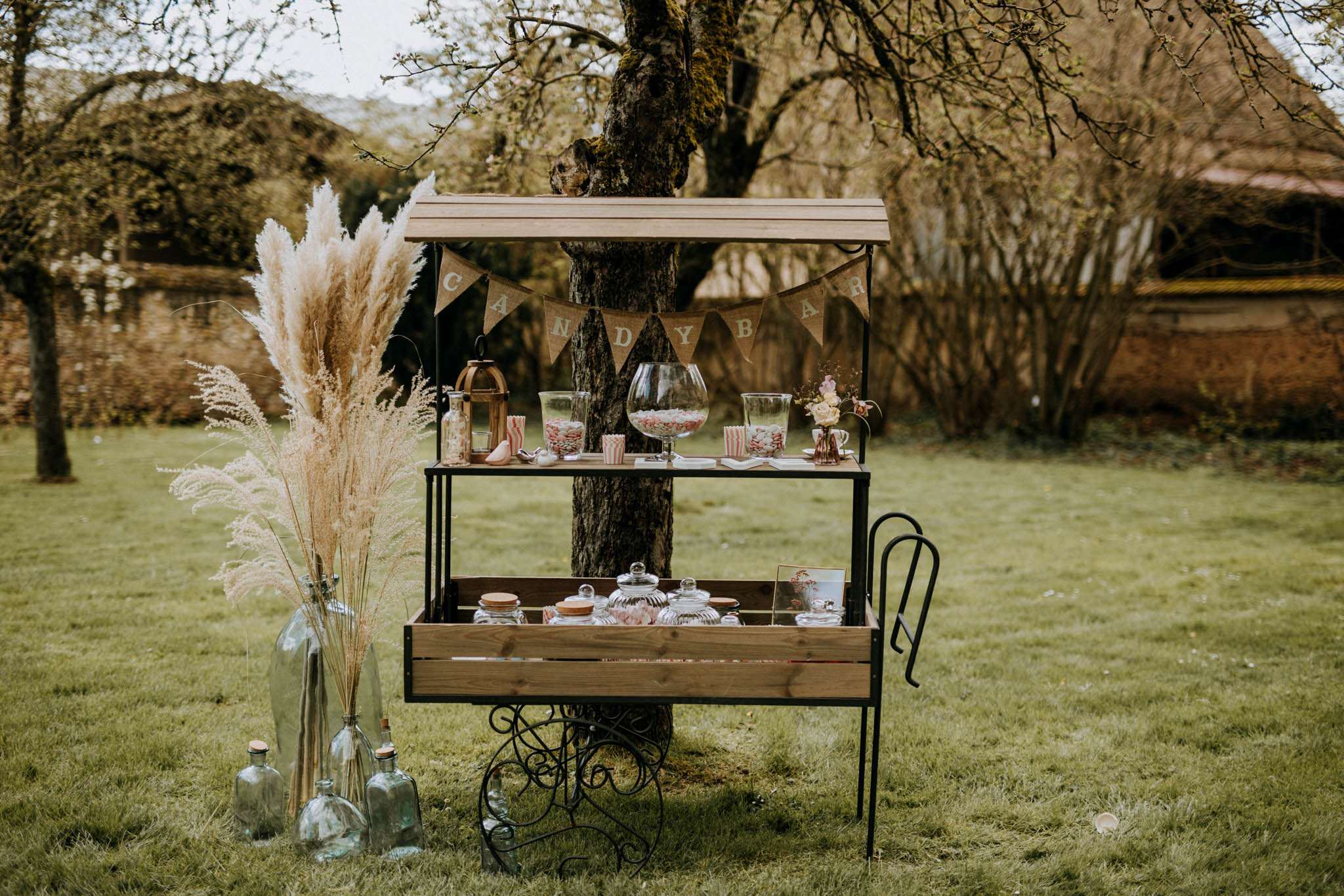 A wedding candy bar station set up outdoors in a garden or grounds, positioned in front of a tree. The setup features a two-tiered wooden cart with a slatted wood roof and black wrought-iron scrolled legs, decorated with a burlap pennant banner spelling out 'CANDY BAR'. The upper tier holds several glass apothecary jars and a large fishbowl filled with pink and white candies, striped paper popcorn cups, a copper lantern, and a small floral arrangement of cream and blush roses. The lower tier contains additional glass jars with cork lids filled with sweets and a small framed photograph. To the left of the cart, large clear glass bottles with cork stoppers are grouped on the ground alongside a tall vase holding dried pampas grass and feathery dried grasses in warm beige tones. The overall styling is rustic-boho with a warm neutral palette. Wide shot, no people visible.