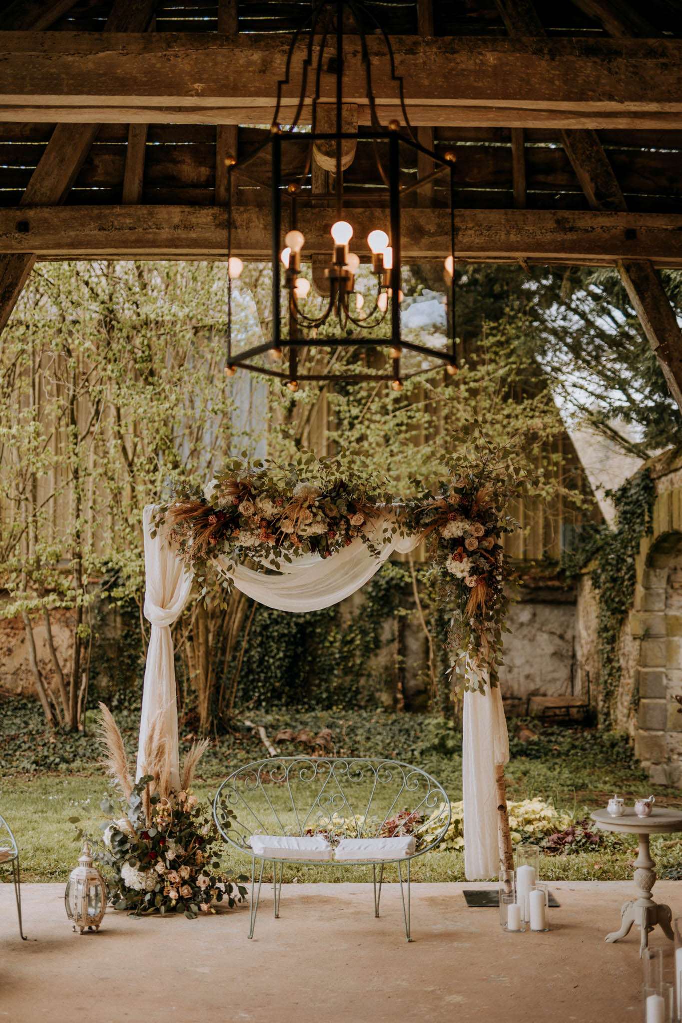 Rustic ceremony setup with chiffon-draped floral arch, pampas grass, wrought-iron loveseat, and Edison bulb chandelier