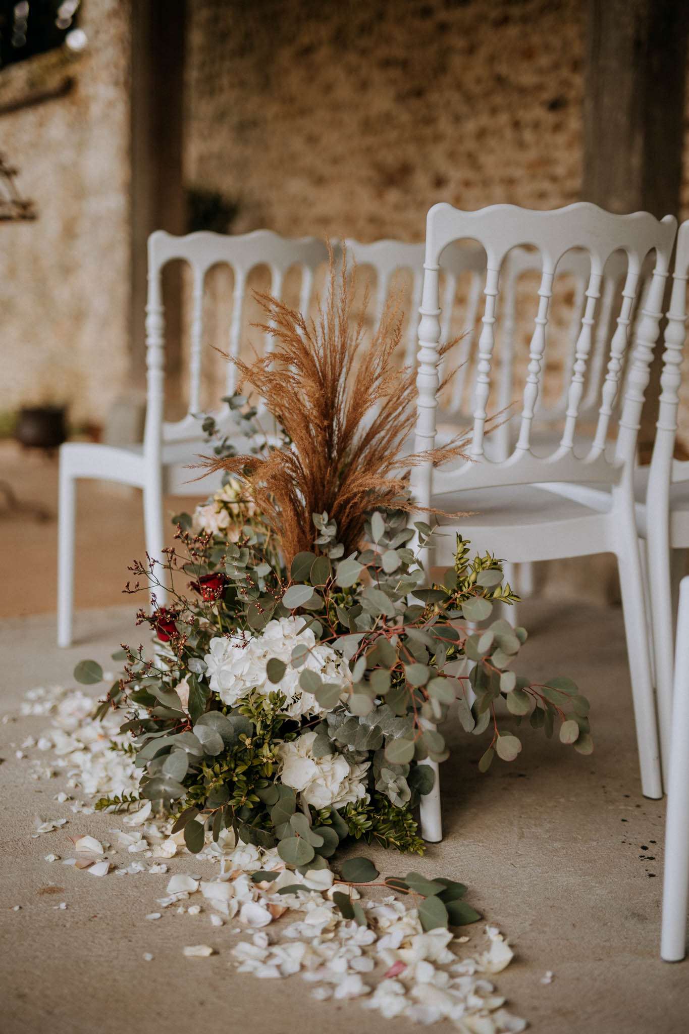 A close-up detail shot of a ceremony aisle floral arrangement placed on the floor beside white Napoleon chairs at an indoor-outdoor venue with exposed stone walls. The arrangement combines white hydrangeas, deep red roses, eucalyptus branches, small-leafed green foliage, dried pampas grass, and burgundy berry stems, creating a boho-rustic aesthetic. Loose white flower petals are scattered across the concrete floor around the base of the arrangement, extending along the aisle. The color palette mixes ivory, deep red, sage green, and warm caramel tones from the dried grasses.