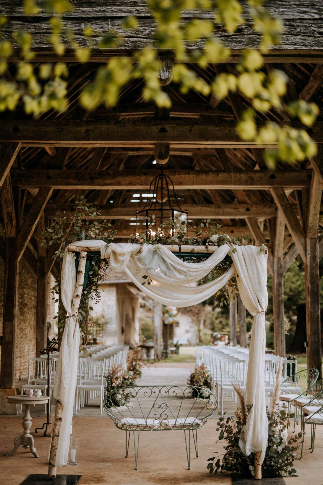 Rustic barn ceremony setup with birch wood arch draped in ivory linen, eucalyptus, and white Chiavari chairs