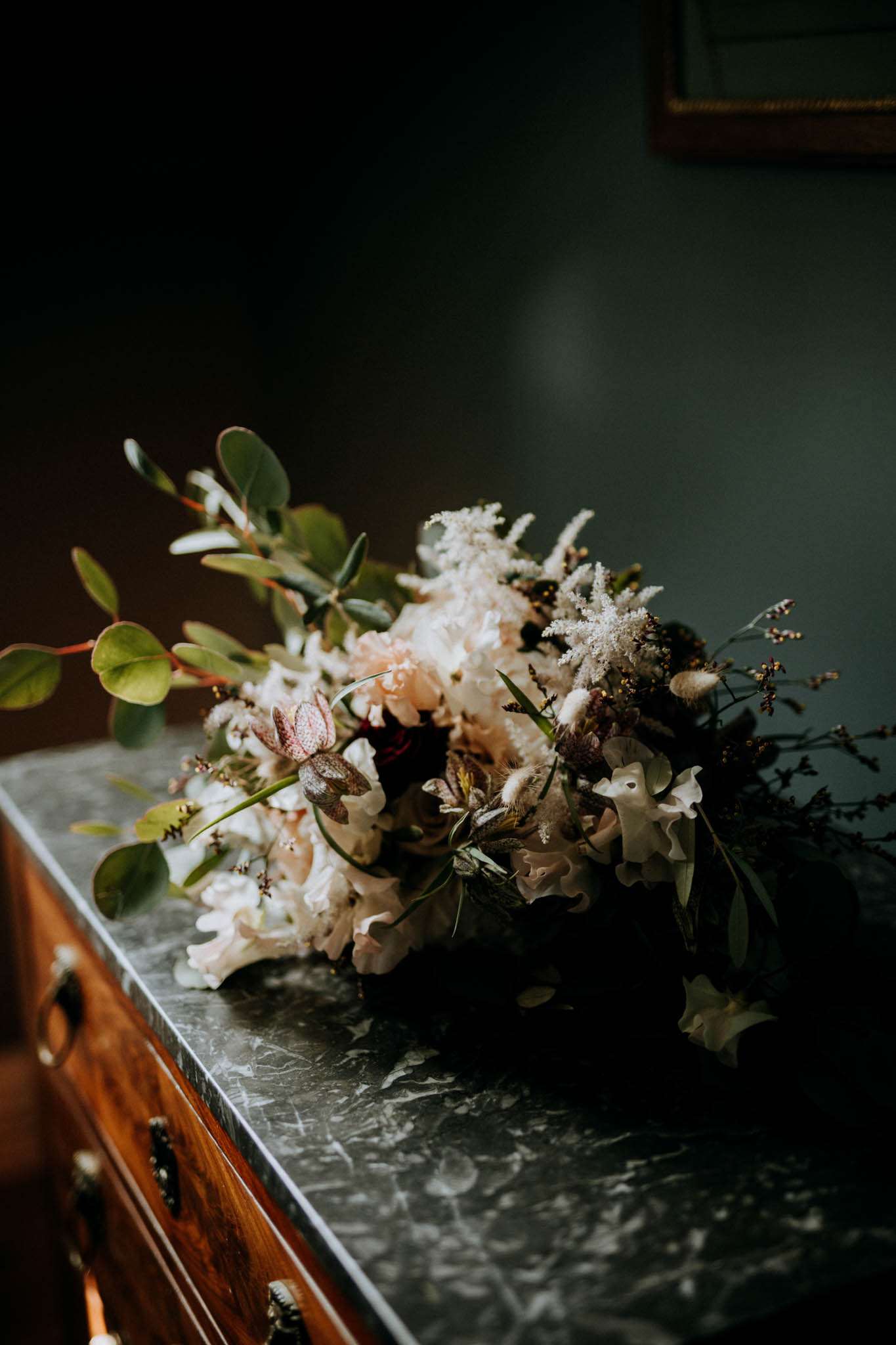 A close-up detail shot of a bridal bouquet resting on a dark grey marble-topped antique wooden dresser with brass hardware. The bouquet is arranged in a loose, garden-style design featuring blush peonies, white astilbe, white sweet peas, burgundy and dark-toned accent blooms, dried bunny tail grasses, small dark berry branches, and trailing eucalyptus foliage. The overall color palette is blush, ivory, deep burgundy, and green. The image is lit with dim, moody natural light against a dark teal-grey wall, with a gold-framed artwork partially visible in the upper right corner, giving the scene a dark, romantic interior atmosphere.