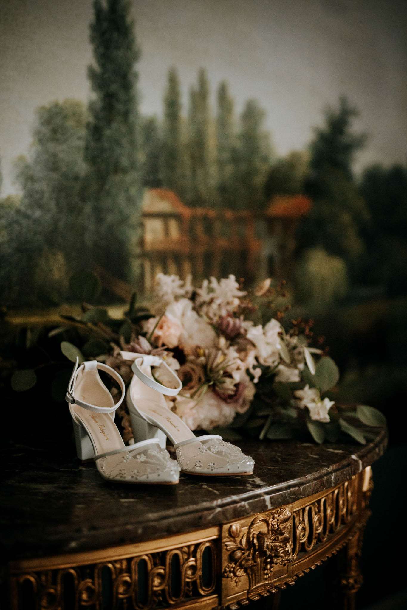 A detail close-up shot of bridal accessories arranged on a dark marble-topped console table with ornate gold carved detailing. A pair of ivory block-heel ankle-strap shoes with embroidered floral toe detailing are placed in front of a loose bridal bouquet featuring blush peonies, mauve and dusty pink blooms, white lily-like flowers, and eucalyptus foliage. The background features a large painted landscape mural depicting trees and a country house, rendered in muted olive and ochre tones. The overall styling suggests a classic French interior setting with a romantic, painterly atmosphere.