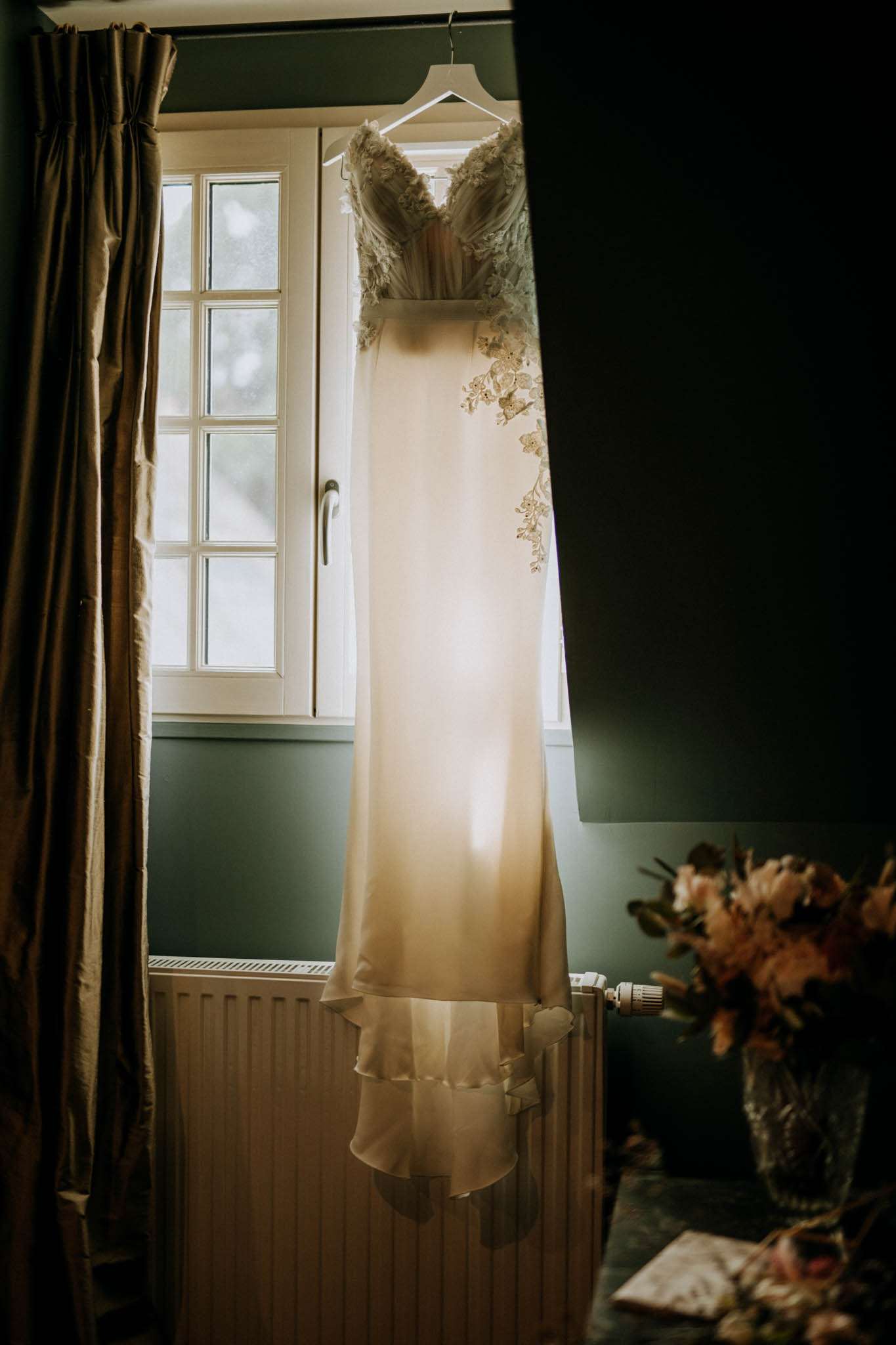 Ivory satin slip gown with floral applique bodice hanging before window in teal-walled room with dried flowers