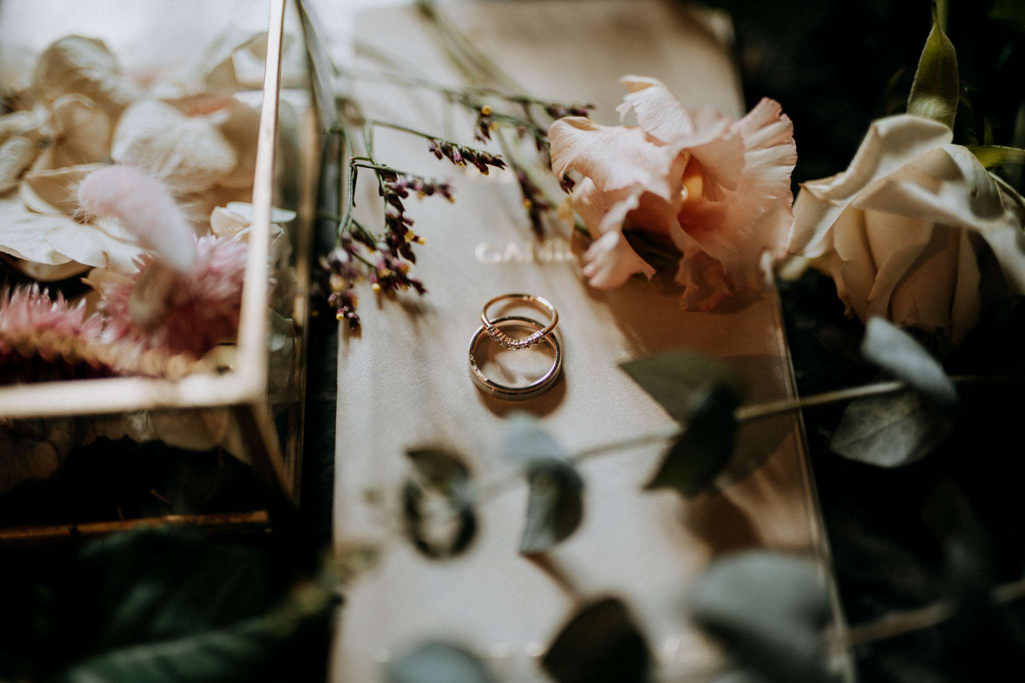 A close-up flat lay detail shot of two wedding rings — one a gold diamond-set band and one a plain gold band — resting on a cream velvet surface, likely a ring box or pouch. The rings are surrounded by blush pink lisianthus blooms, dried purple statice sprigs, eucalyptus leaves, and soft pink dried pampas-style florals. To the left, a brass and glass geometric terrarium ring box holds additional dried and fresh florals in blush and cream tones. The overall styling palette is blush pink, cream, and muted sage, consistent with a romantic boho aesthetic. Soft, moody natural lighting with shallow depth of field.