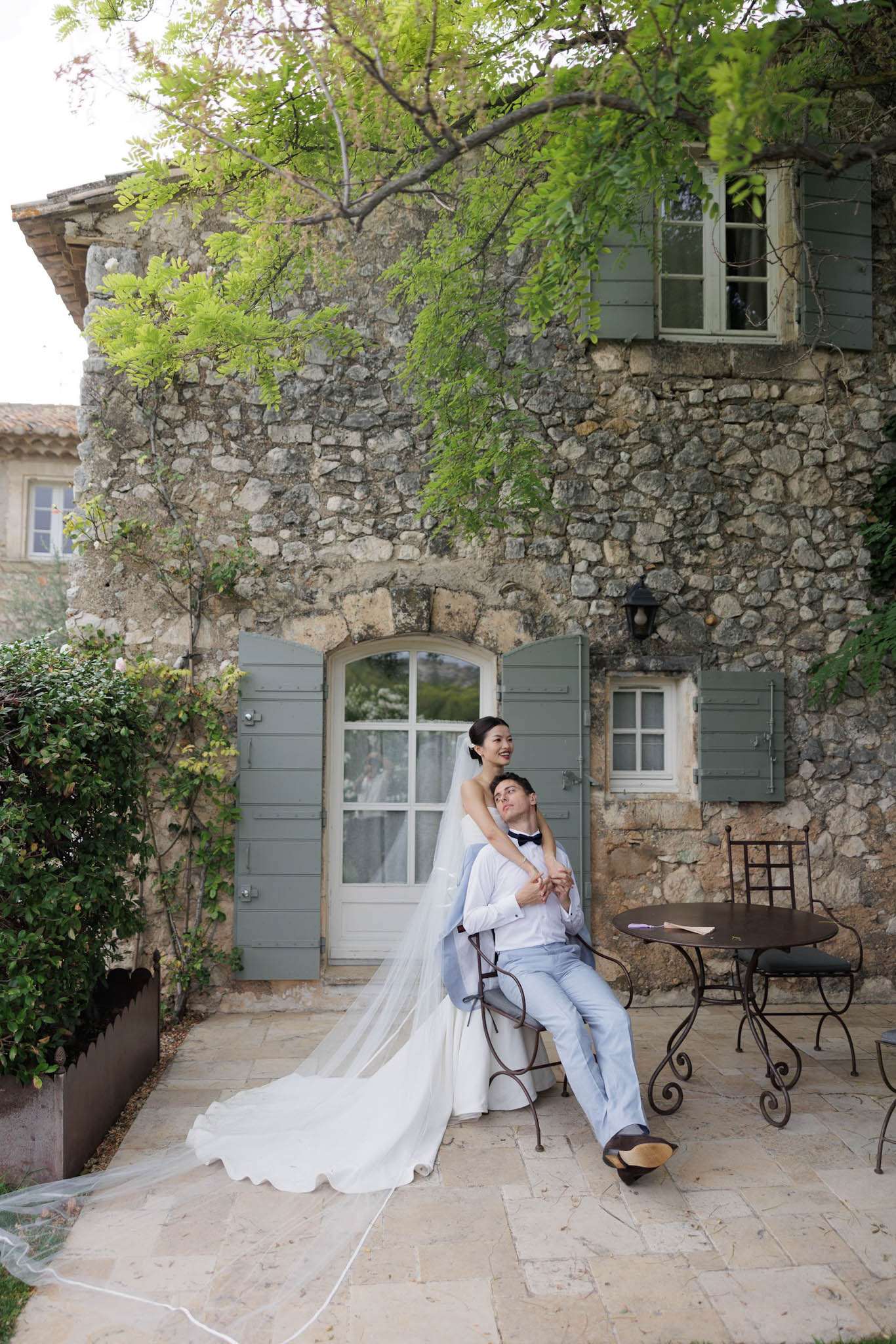 A couple portrait taken outdoors on a stone-paved terrace in front of a two-storey Provençal stone building with sage green shutters and an arched doorway. The groom is seated on a wrought iron chair wearing light blue trousers, a white shirt, and a black bow tie, with a light blue suit jacket draped over the chair back; the bride stands behind him with her arms around his shoulders, wearing a white strapless gown with a long cathedral-length veil that fans across the terrace floor. Both are looking upward and smiling. A small round wrought iron bistro table and additional chairs are visible to the right. The styling is classic and relaxed, combining a formal white bridal gown with a more casual light blue groom's ensemble. The shot is a full-length portrait taken from a slight distance, capturing both figures and the full architectural facade of the building.