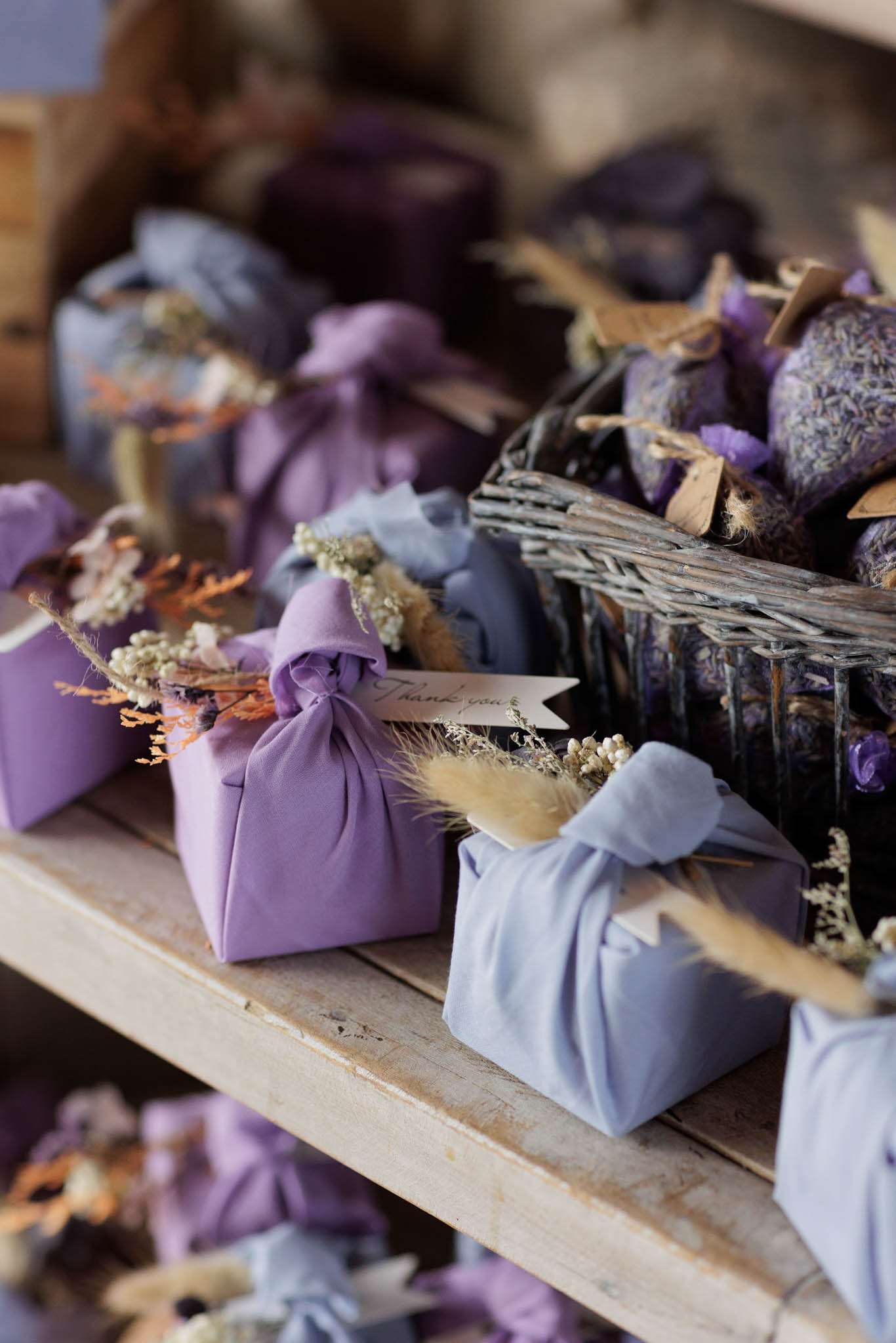 Close-up detail shot of wedding guest favors displayed on a rustic wooden shelf. The favors are wrapped in the furoshiki style using fabric in two tones — medium purple and dusty periwinkle blue — each tied at the top and decorated with small bunches of dried botanicals including pampas grass, orange dried stems, cream statice, and dried lavender. One purple favor features a small calligraphed 'Thank you' gift tag. A wicker basket in the background holds additional lavender-filled sachets in matching purple fabric. The overall decor palette of purple, periwinkle, and dried natural tones suggests a rustic-romantic or Provençal styling theme.