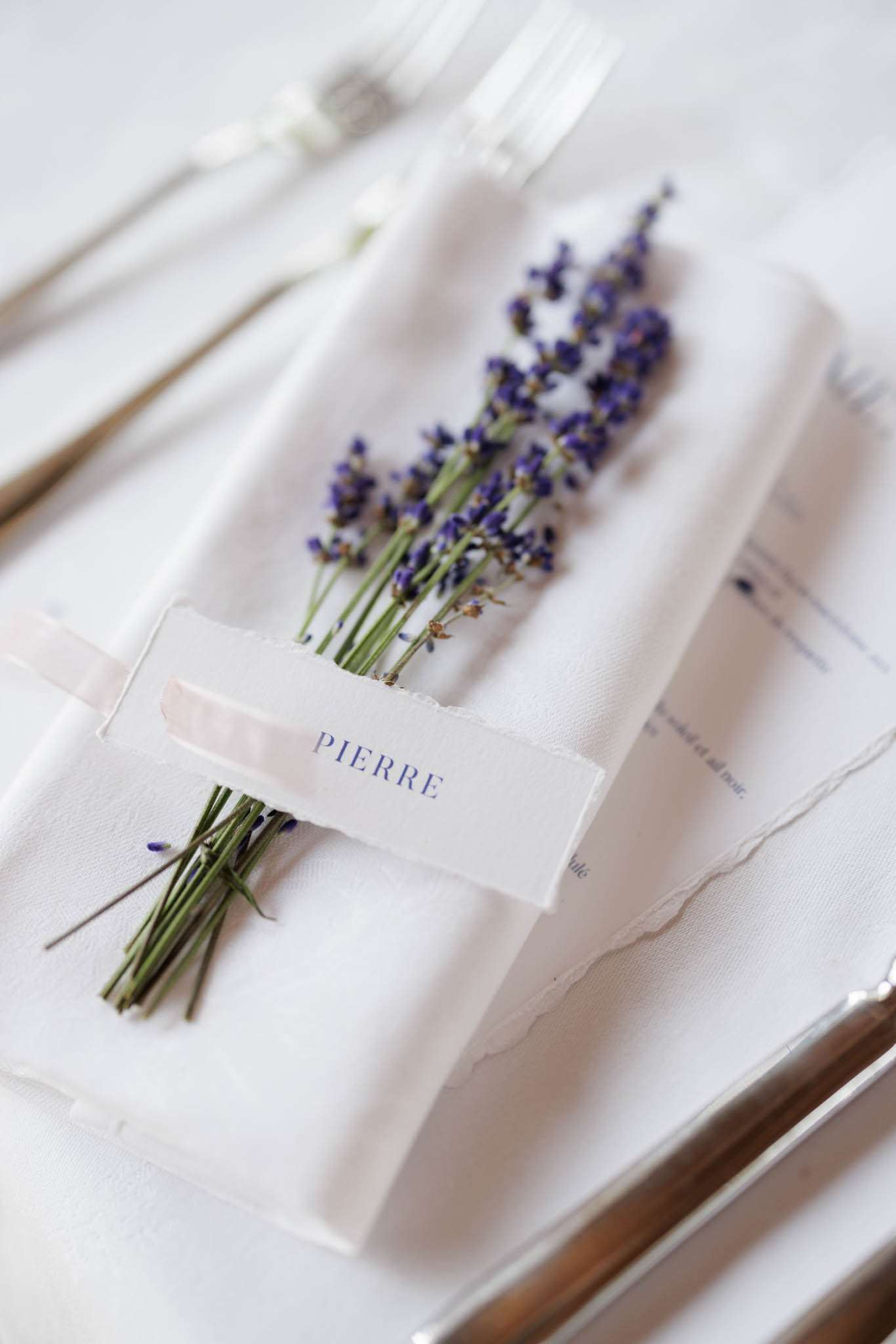 Place setting with white napkin tied with fresh lavender, deckle-edged place card reading Pierre, and gold flatware