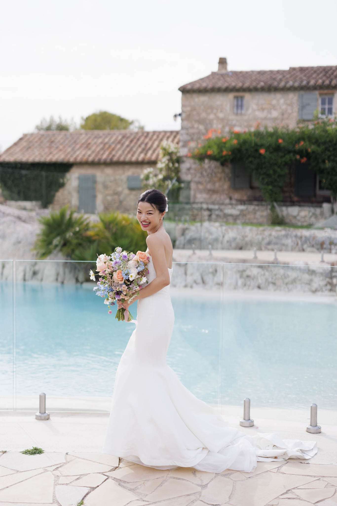 Bride in strapless white mermaid gown holding peach and lavender bouquet on stone pool terrace at Provencal venue