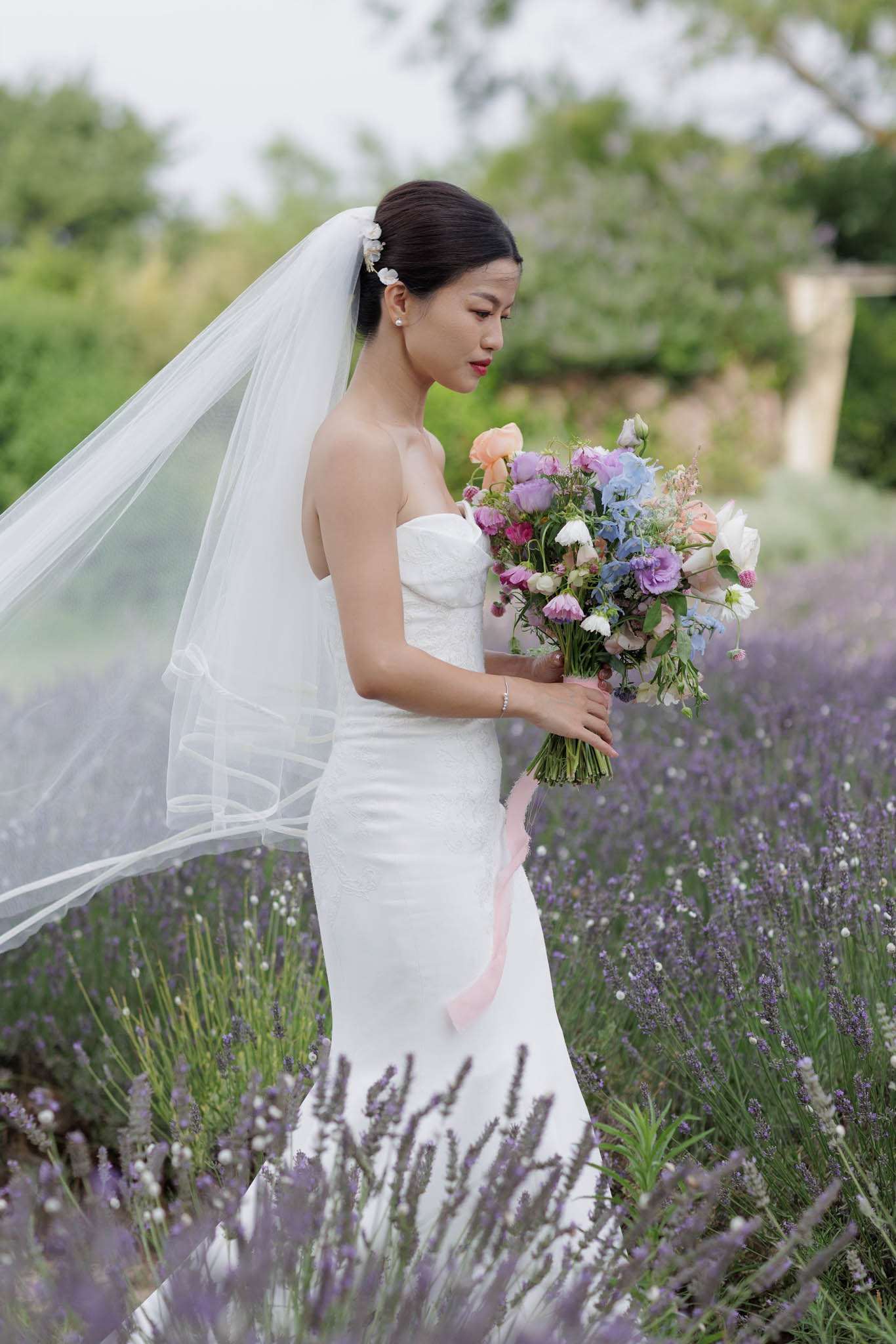 Bride in profile in lavender field holding multicolored bouquet of roses, hydrangea, and lisianthus