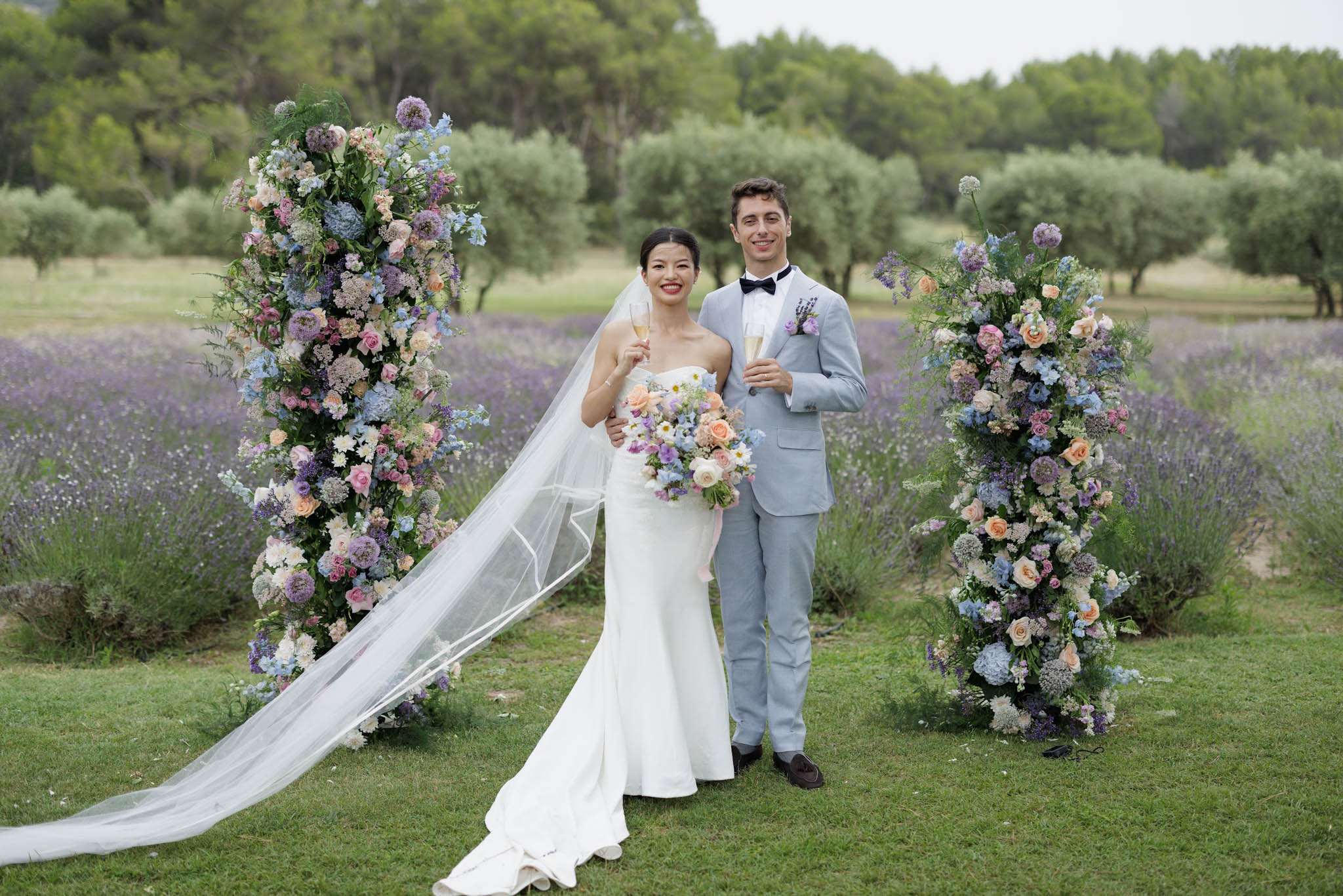 Bride and groom hold champagne flutes between floral columns in a Provencal lavender field