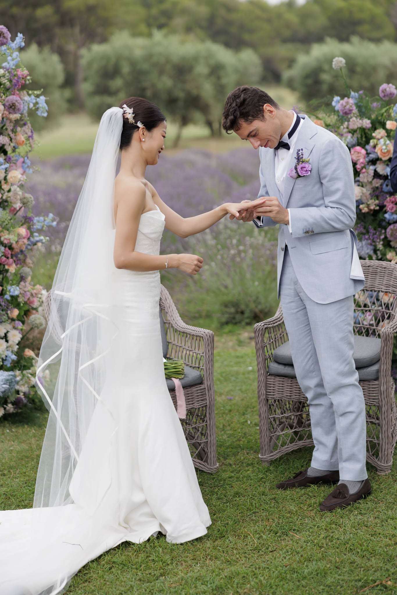 The ring exchange moment of an outdoor ceremony taking place in what appears to be a Provençal lavender field, with rows of purple lavender visible in the background. The bride wears a strapless white fitted gown with a long cathedral veil edged in ribbon and a floral hair accessory, while the groom places a ring on her finger; he is dressed in a light blue suit with a navy bow tie and a boutonnière of purple lisianthus and peach roses. Two large floral arch columns flank the couple, composed of blue delphinium, blush and peach roses, purple lisianthus, and allium in a pastel palette. The couple stands on a lawn between two grey wicker ceremony chairs, and the composition is a medium portrait shot capturing both figures from head to toe with the lavender field softly blurred behind them.