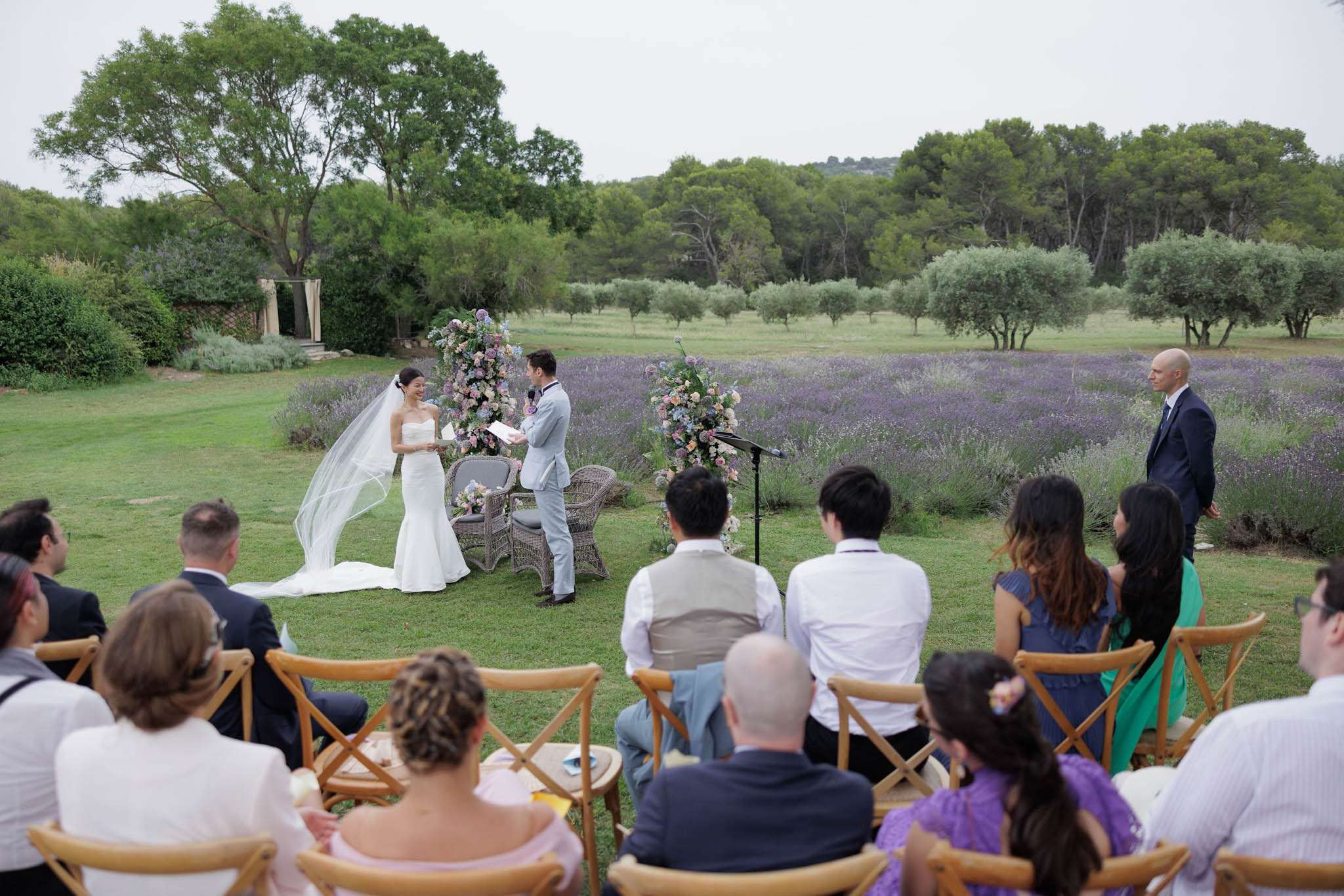 Outdoor wedding ceremony on lawn before lavender field with pastel floral columns and guests in wooden chairs