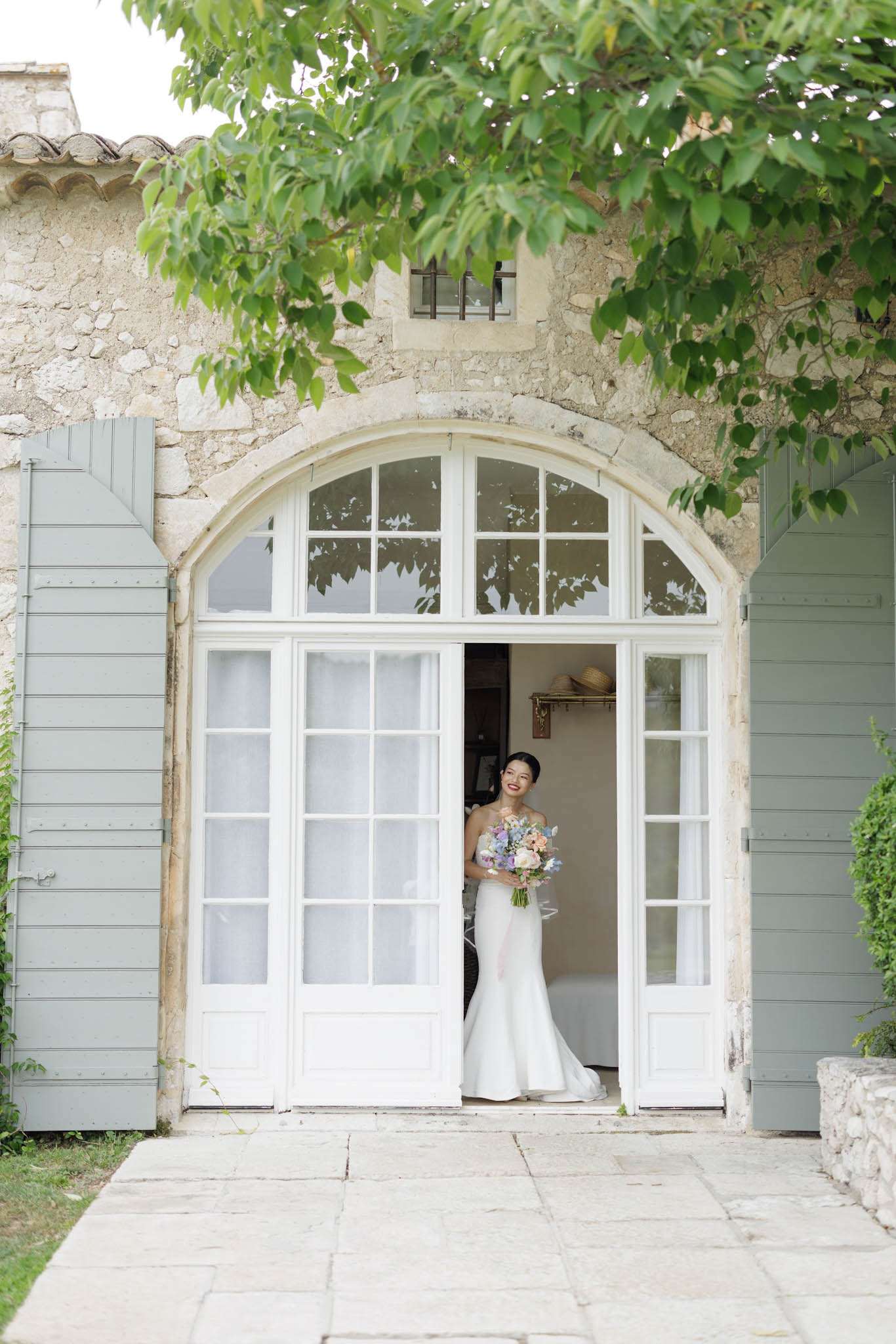A bride stands in the open doorway of a French stone building, captured in a medium portrait shot. She is wearing a fitted strapless ivory mermaid-style gown and holds a loose, garden-style bouquet featuring soft lavender, blush, and white blooms including what appear to be sweet peas, ranunculus, and delicate wildflowers. The building facade features a large arched white French door with divided-light panes and sage green shutters on either side, characteristic of a Provençal mas or château outbuilding. The overall styling is classic with a soft, romantic floral palette. Potential venue feature image.