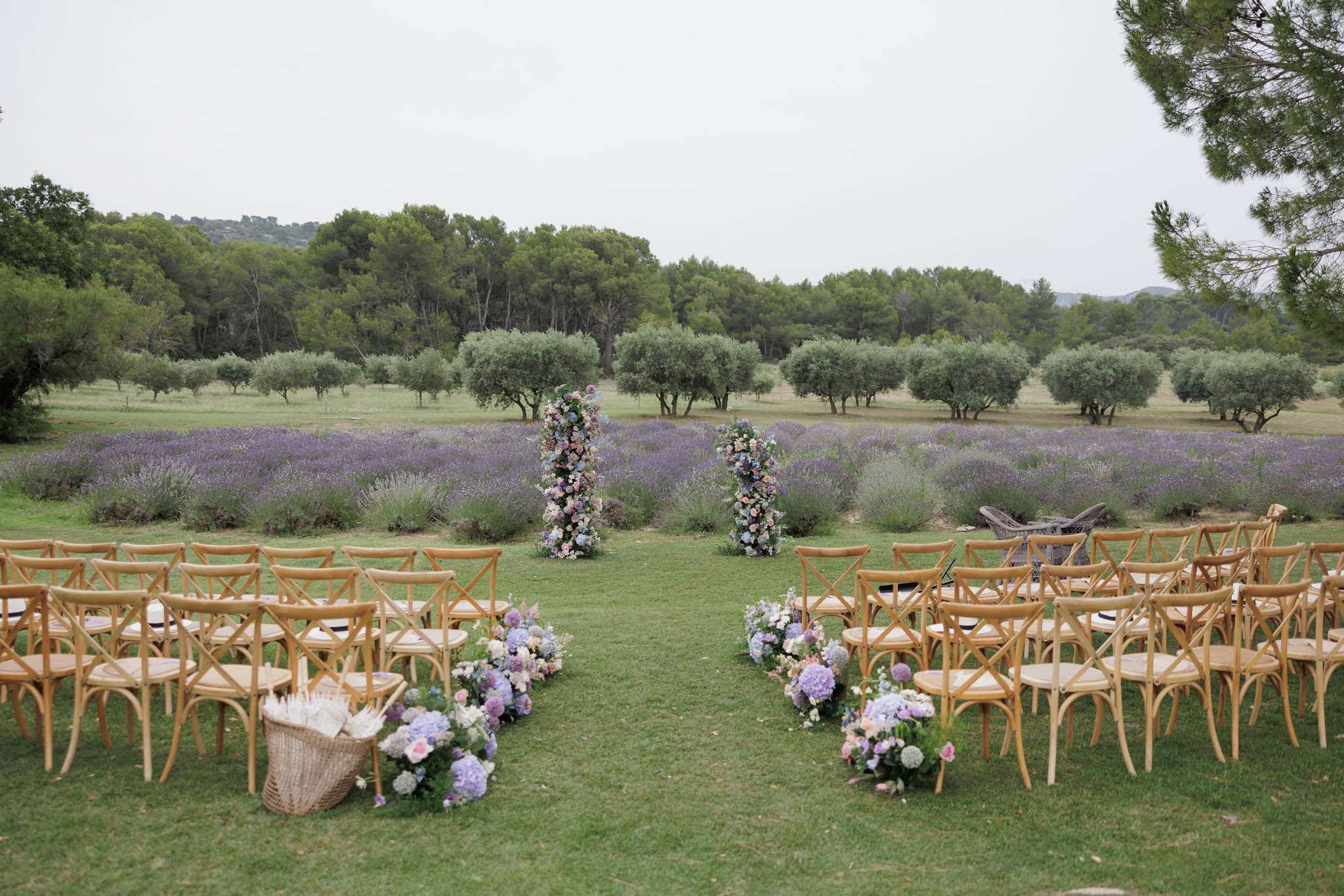 An outdoor wedding ceremony setup on a lawn, photographed before guests arrive, in a wide shot that captures the full layout. Rows of natural wood cross-back chairs are arranged on either side of a grass aisle, with a wicker basket holding white paper fans placed at the end of one row. The aisle is lined with low floral arrangements of lavender-blue hydrangeas, blush pink roses, and white blooms placed directly on the ground. At the altar position, two tall freestanding floral columns feature a pastel mix of pink, blush, white, and lilac florals. The ceremony faces out toward rows of olive trees and a lavender field in full purple bloom, creating a distinctly Provençal setting. The overall decor palette is soft pastel with a garden-romantic style.