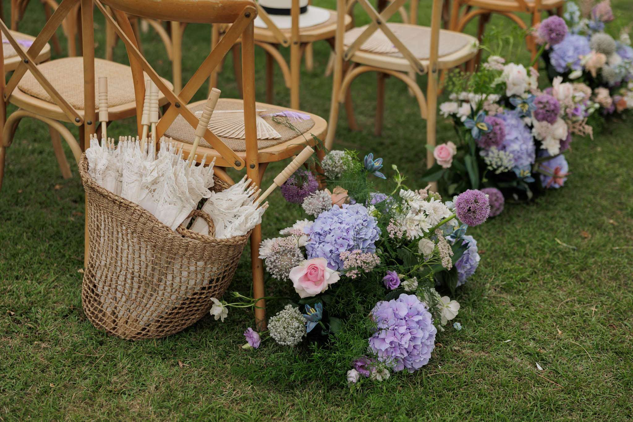 Aisle floral arrangements with purple hydrangeas, pink roses, and alliums beside cross-back chairs and parasol basket
