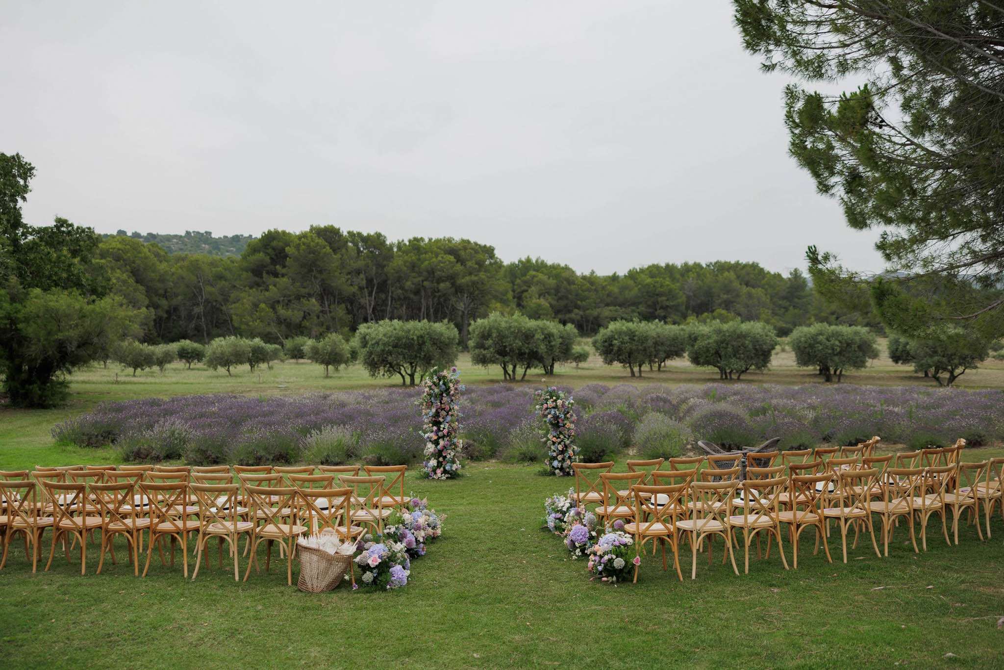 An outdoor wedding ceremony setup photographed before guests arrive, positioned on a lawn directly in front of rows of blooming lavender fields with an olive grove beyond. Natural wood cross-back chairs are arranged in two sections separated by a central aisle, with approximately eight rows on each side. At the front of the aisle, two tall floral columns decorated with pastel pink, peach, and lilac blooms mark the ceremony space. Clusters of hydrangeas, roses, and mixed pastel florals are placed at the base of the first chairs on each side of the aisle, and a wicker basket sits at the end of one row. The overall decor palette is soft pastel — lavender, blush pink, and peach — with a rustic-romantic styling. Wide shot taken from behind the seating area looking toward the lavender field and treeline. Potential venue feature image.