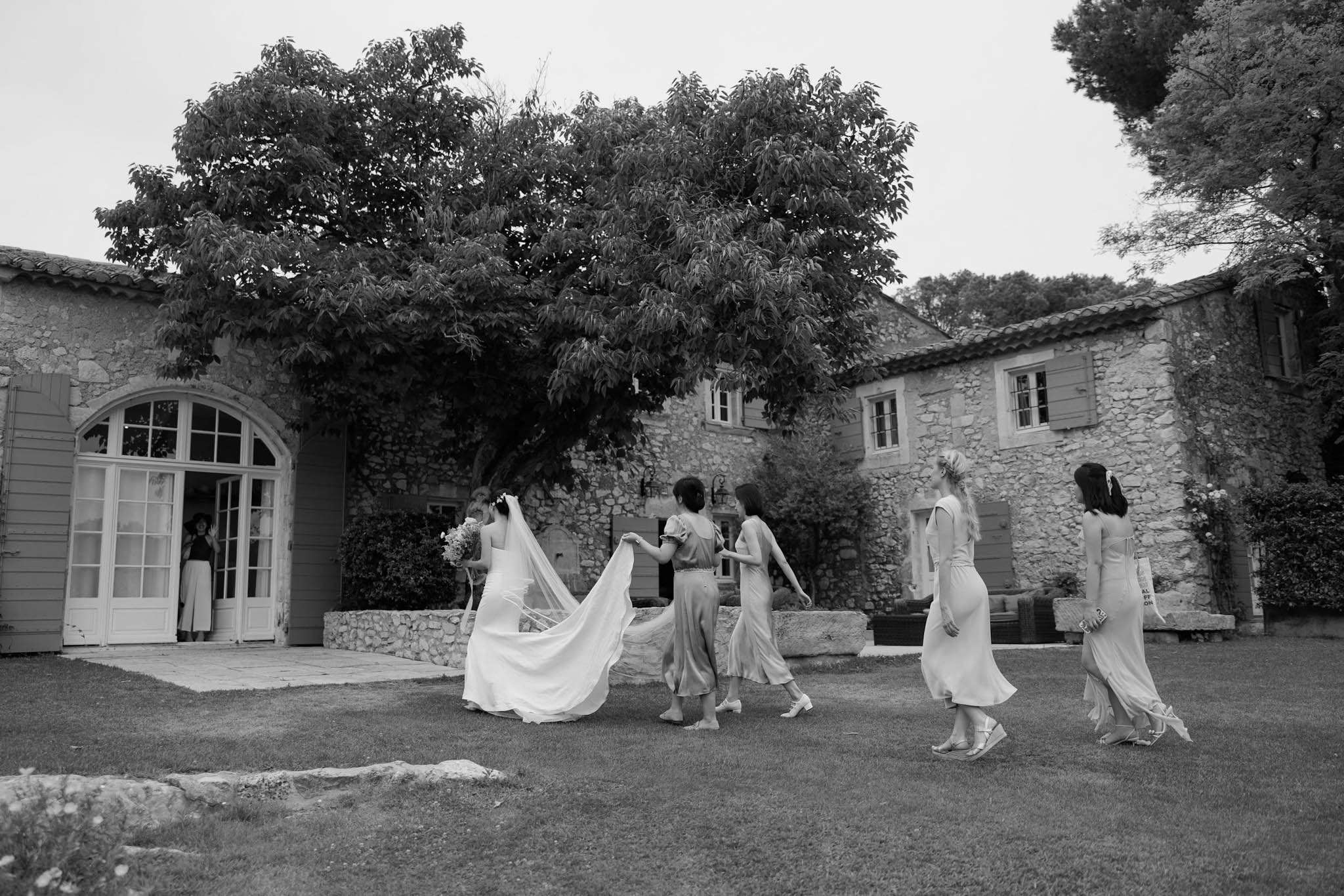 Black-and-white shot of a bride walking toward a Provencal stone mas as four bridesmaids carry her cathedral veil