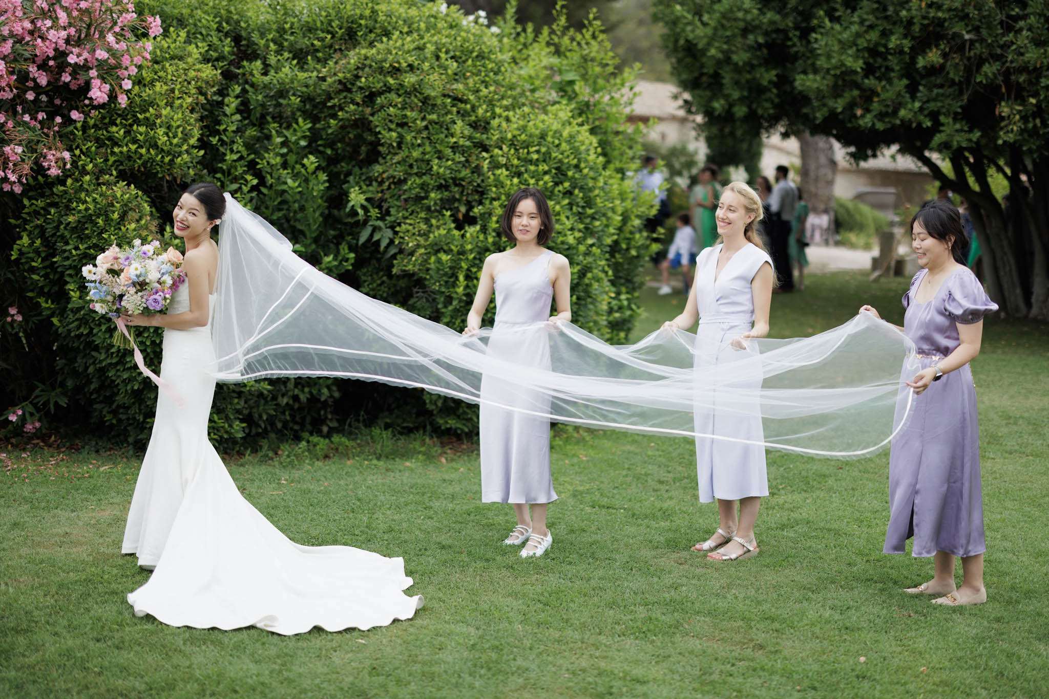 Bride looks over her shoulder as three bridesmaids in mismatched lavender outfits hold her cathedral veil on a lawn