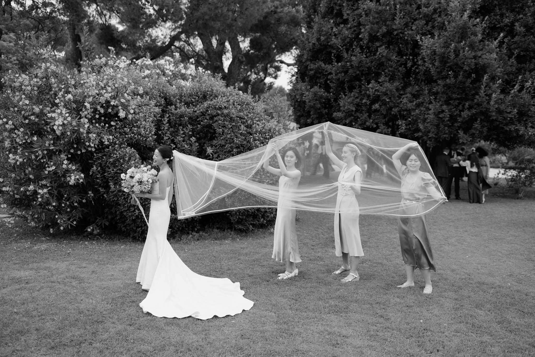This black-and-white outdoor portrait captures a bride and three bridesmaids on a lawn, with a small group of guests visible in the background. The bride stands on the left facing away from camera, wearing a fitted sleeveless mermaid-style gown with a train and holding a round bouquet; a long cathedral-length veil with an edge trim billows outward as the three bridesmaids lift and spread it dramatically above their heads. The bridesmaids wear midi-length dresses in varied styles — the tones suggest satin or silk fabrics — and flat or low-heeled sandals, each in a slightly different silhouette. The image is shot in high contrast black and white with strong mid-tones, rendered as a wide environmental portrait that emphasizes the veil's full spread against the garden setting.
