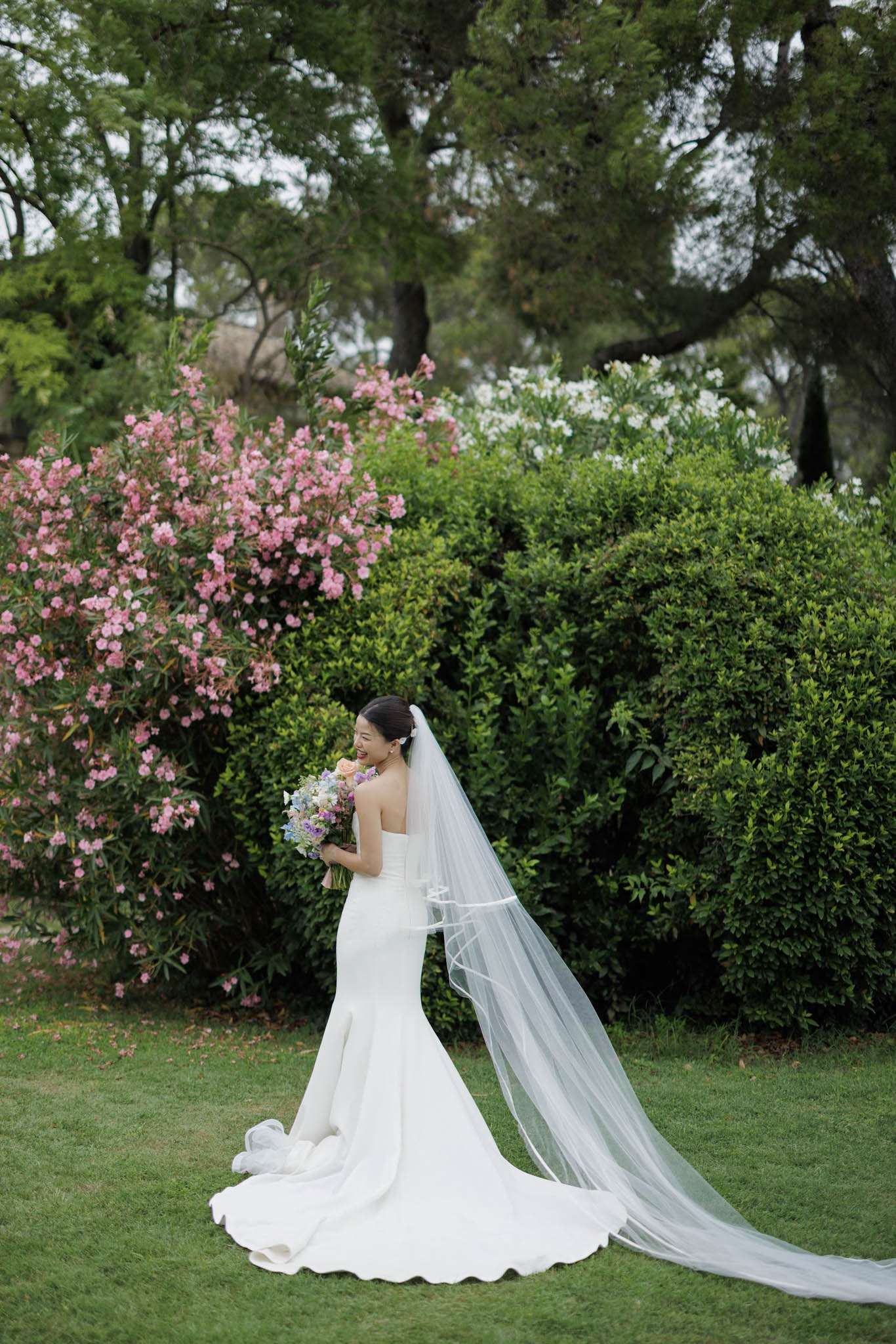 A bridal portrait taken outdoors in a garden setting, with the bride standing on a lawn and turning slightly to one side while laughing. She wears a fitted ivory strapless mermaid-style gown with a long train and a cathedral-length plain-edge tulle veil that trails across the grass. She holds a loosely arranged bouquet featuring lavender, blush, and pale blue flowers. Her dark hair is styled in a low updo with a small floral hair accessory. The composition is a full-length portrait shot from a medium distance, capturing the full veil and train spread across the lawn.