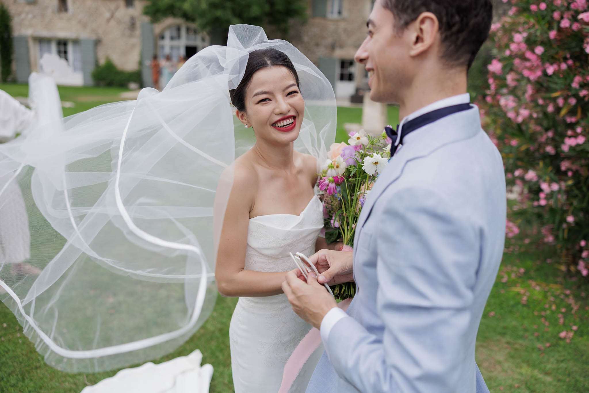 Bride and groom exchanging rings during outdoor chateau ceremony, bride in white gown with flowing veil and garden bouquet
