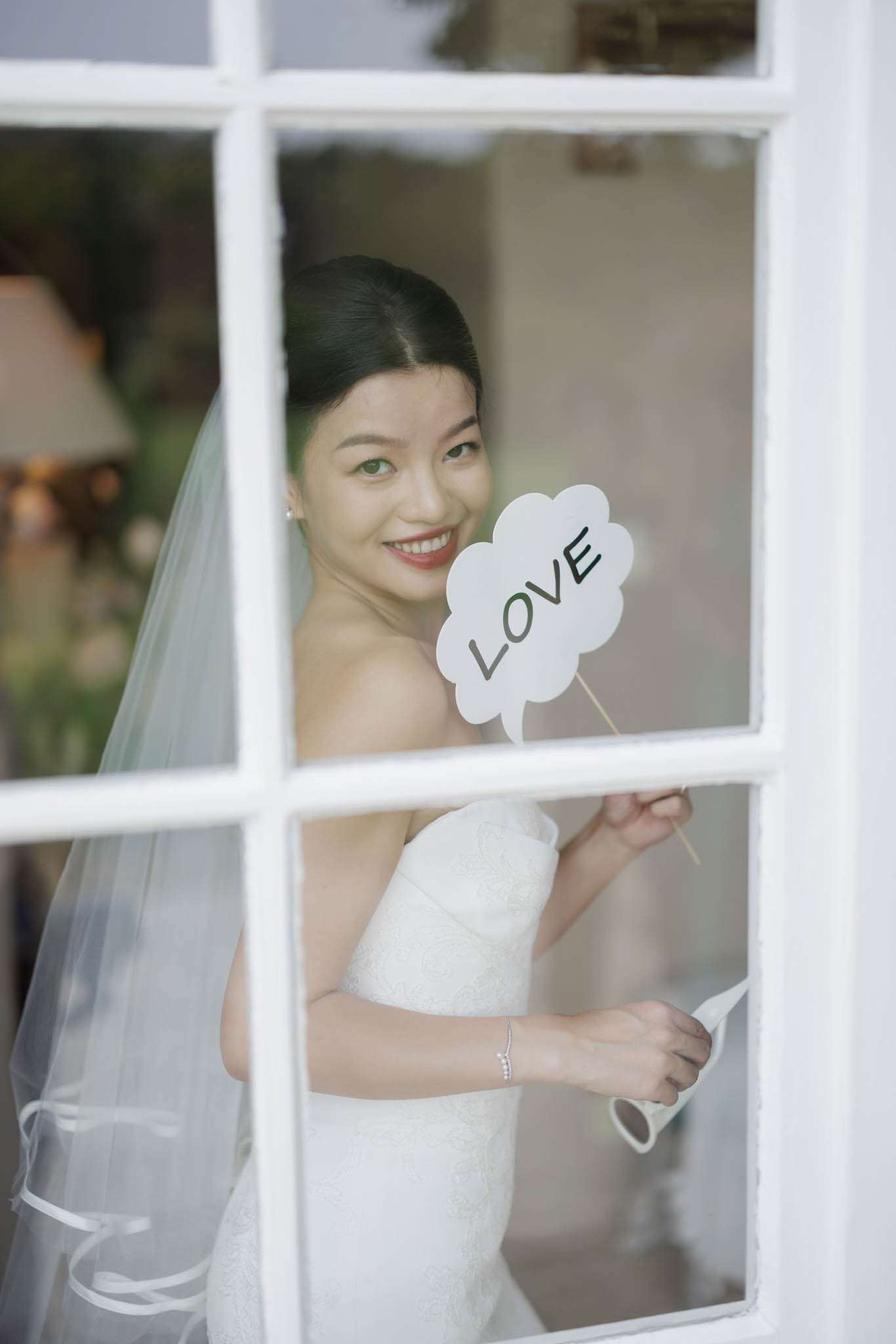 A bride poses indoors behind a white-framed multi-pane window, photographed through the glass from outside. She is wearing a white strapless fitted gown with a subtle texture, a long cathedral-length veil with a satin ribbon edge, and a delicate diamond line bracelet. Her dark hair is styled in a sleek low bun. She holds a white cloud-shaped photo booth prop on a wooden stick printed with the word 'LOVE' in dark lettering, and smiles directly at the camera. The composition is a portrait shot framed by the window muntins, with a warmly lit interior visible softly in the background.