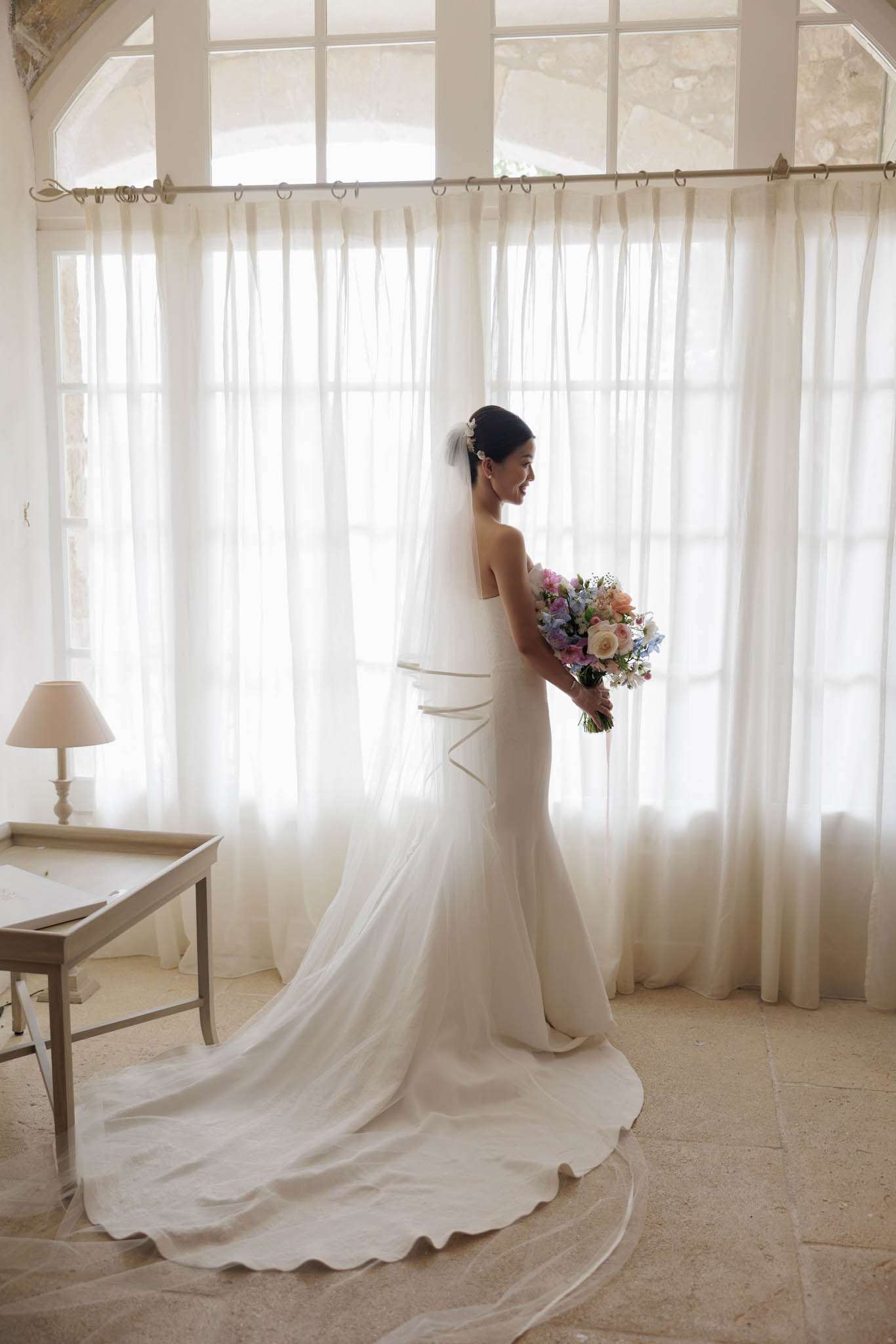A bridal portrait taken indoors in a bright, high-ceilinged room with tall arched windows dressed in sheer ivory linen curtains, likely within a French château or historic stone building. The bride stands in three-quarter profile facing right, wearing a fitted ivory strapless gown with a structured corseted back detail and a long cathedral-length train pooling on the stone tile floor, paired with a long plain-edge veil secured with a small floral hair accessory. She holds a loose, garden-style bouquet composed of blush peach roses, soft blue delphinium, lilac sweet peas, and small white filler flowers. Natural backlight from the windows creates a soft, bright exposure, and a small table lamp and writing desk are visible to the left. Full-length portrait composition with a classic, clean aesthetic.