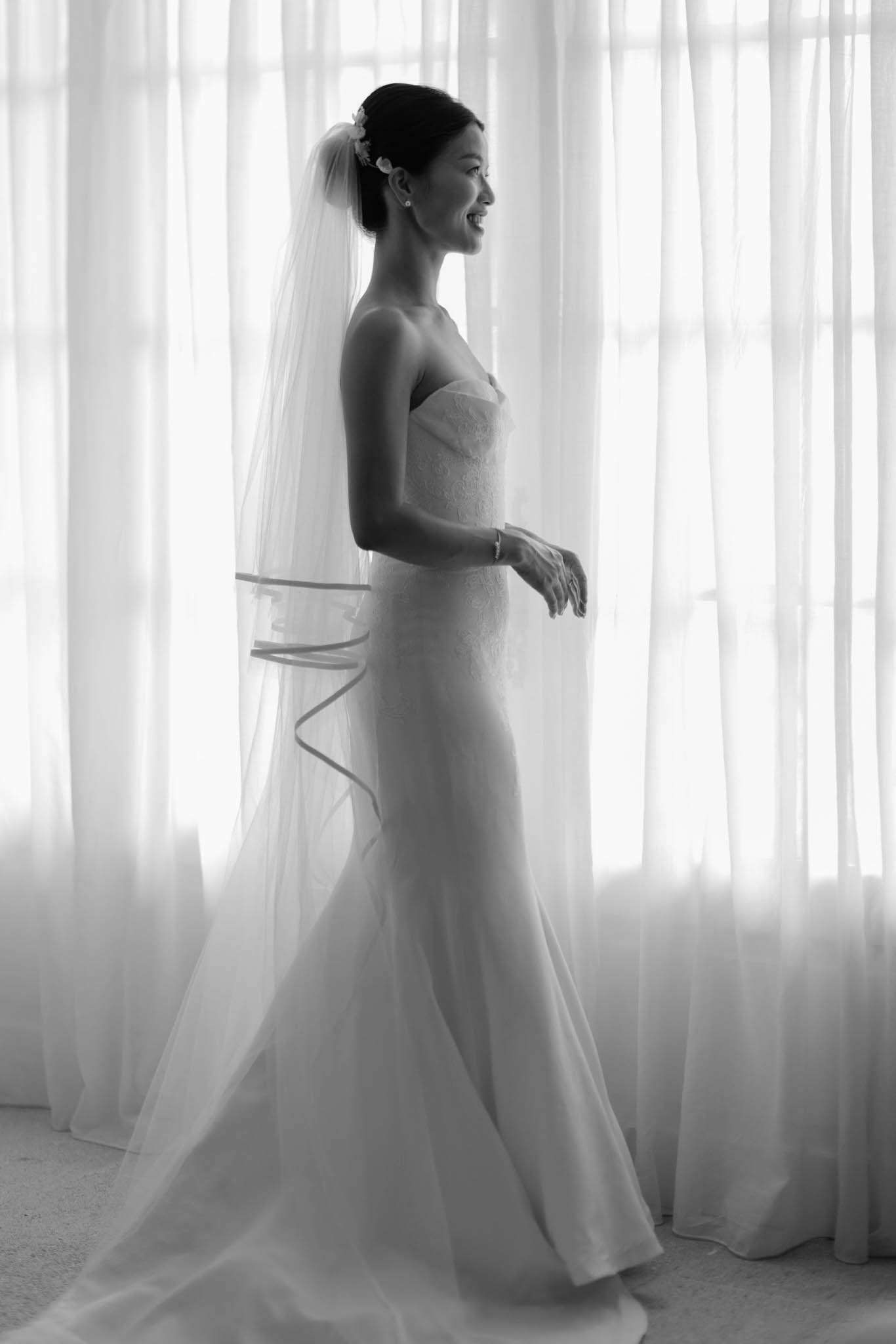 Black and white bride in strapless lace gown with cathedral veil and ribbon edge before sheer curtains