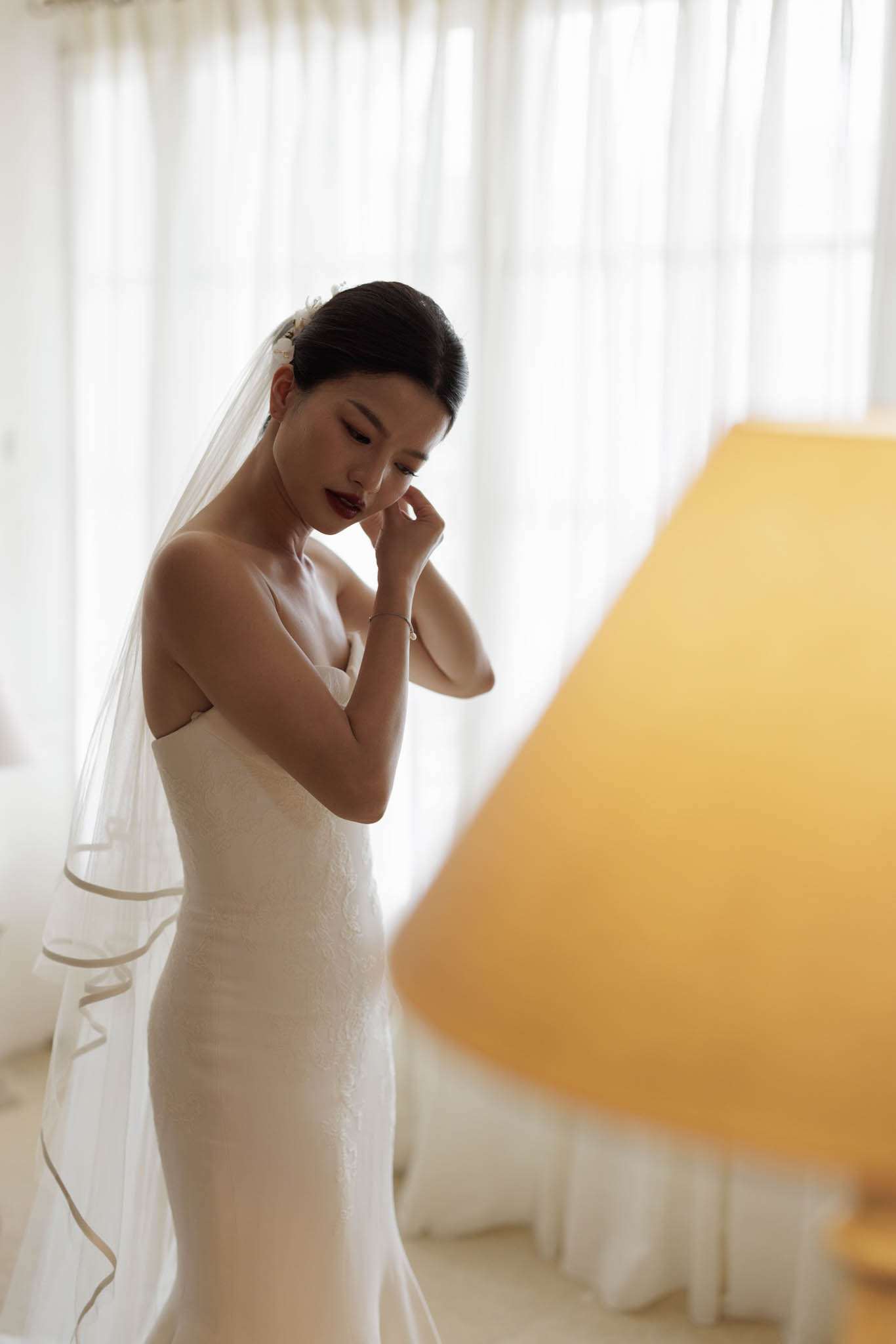 A bride is photographed indoors during the getting-ready portion of her wedding day, standing near a sheer white curtained window that provides soft natural backlighting. She wears a fitted, strapless ivory crepe or lace-textured gown with a sleek silhouette, paired with a cathedral-length veil trimmed with a satin ribbon edge and small white floral hair accessories at the crown. Her dark hair is pulled back in a low, smooth updo, and she wears deep berry-toned lip color with a delicate pearl or chain bracelet on her wrist. She appears to be adjusting her earring, with her gaze downward. In the right foreground, a large yellow lampshade is partially visible and softly out of focus, adding a warm amber tone to the composition. The shot is a close-to-mid portrait with a shallow depth of field, styled with a clean, modern aesthetic.