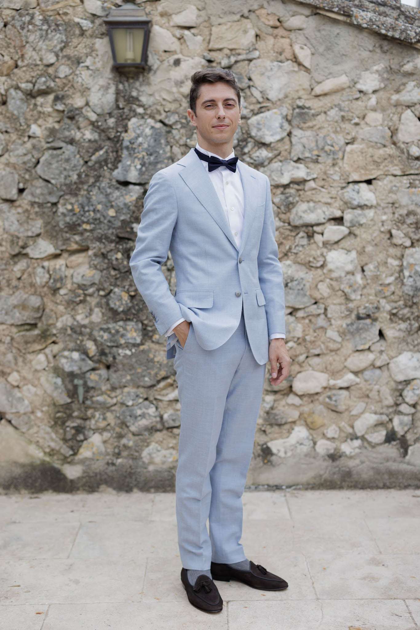 A groom stands alone for a portrait outdoors against a rustic stone wall with a wall-mounted lantern visible above. He is wearing a light blue two-piece suit with a white dress shirt, a navy blue bow tie, and dark brown tassel loafers. One hand is tucked casually into his trouser pocket. The shot is a full-length portrait taken at eye level, with the groom centered in the frame on a stone-paved surface. The overall styling aesthetic is classic with a relaxed, summery feel.