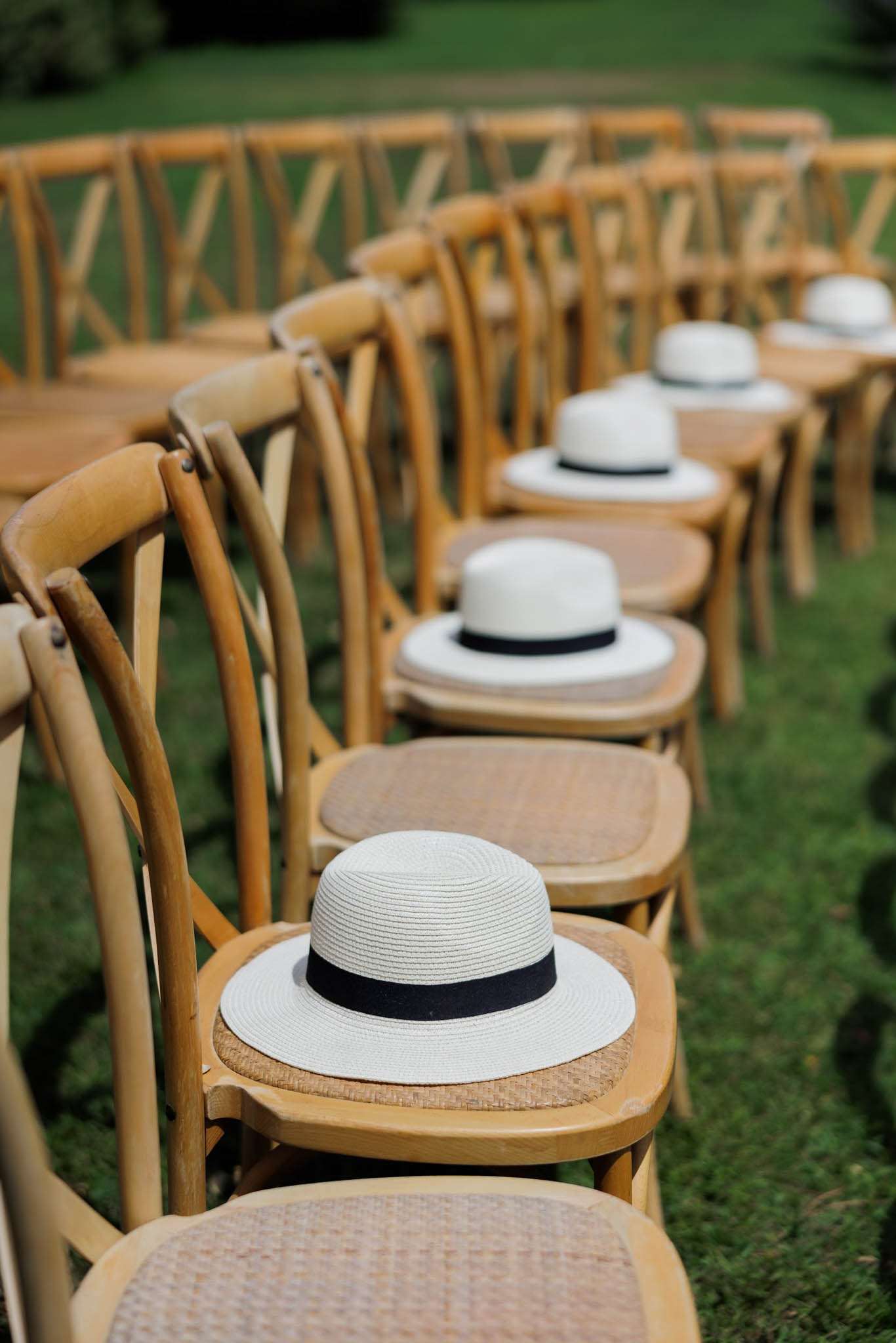 An outdoor ceremony setup showing rows of natural wood cross-back chairs arranged on a lawn, with white straw boater hats trimmed with a black grosgrain ribbon placed on the seats as guest favors or sun protection gifts. The hats are positioned on one side of the aisle, visible along at least four to five chairs receding into the background with shallow depth of field. The styling is classic and clean, with the black-and-white hat detail contrasting against the warm honey-toned wood of the chairs. This is a detail shot taken from a low angle along the aisle.