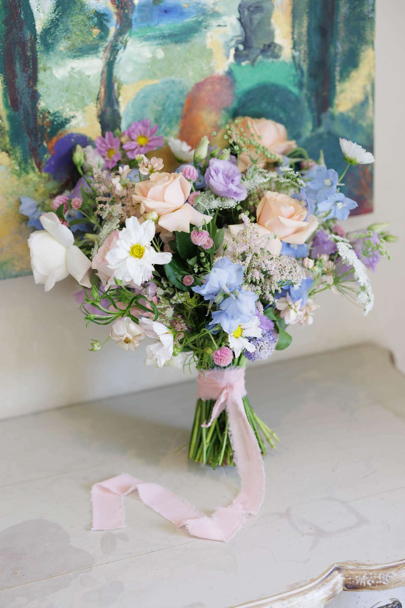 Garden-style bouquet of peach roses, blue delphiniums, and mauve asters with blush ribbon on marble