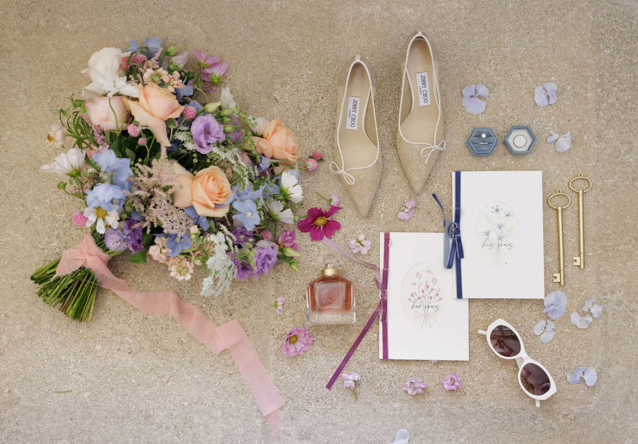 A bridal flat lay detail shot arranged on a sandy stone surface, featuring a loose garden-style bouquet with peach roses, blue delphinium, purple lisianthus, white cosmos, pink wildflowers, and mixed greenery, wrapped with a blush pink silk ribbon. Surrounding the bouquet are a pair of champagne gold pointed-toe Jimmy Choo flats with bow detail, two hexagonal dusty blue velvet ring boxes containing an engagement ring and a wedding band, two white vow booklets labeled 'her vows' and 'his vows' with watercolor floral illustrations tied with burgundy and navy ribbon, a Guerlain perfume bottle, two gold skeleton keys, white cat-eye sunglasses, and scattered loose purple petals and pink cosmos blooms. The overall color palette is soft and romantic, mixing peach, lavender, dusty blue, and burgundy across florals and stationery.
