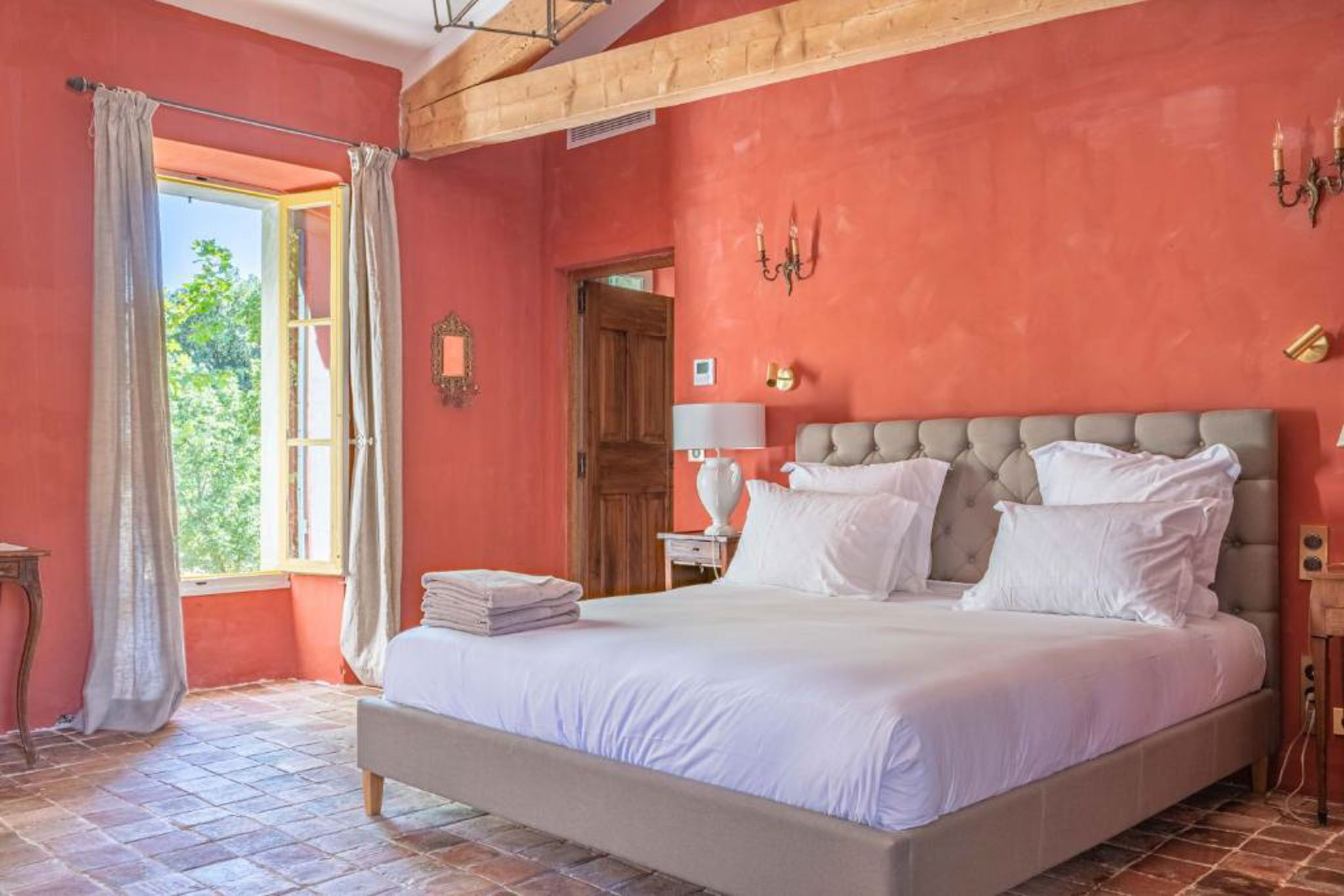Guest bedroom with terracotta walls, exposed wooden beams, king bed with grey headboard, and open French window