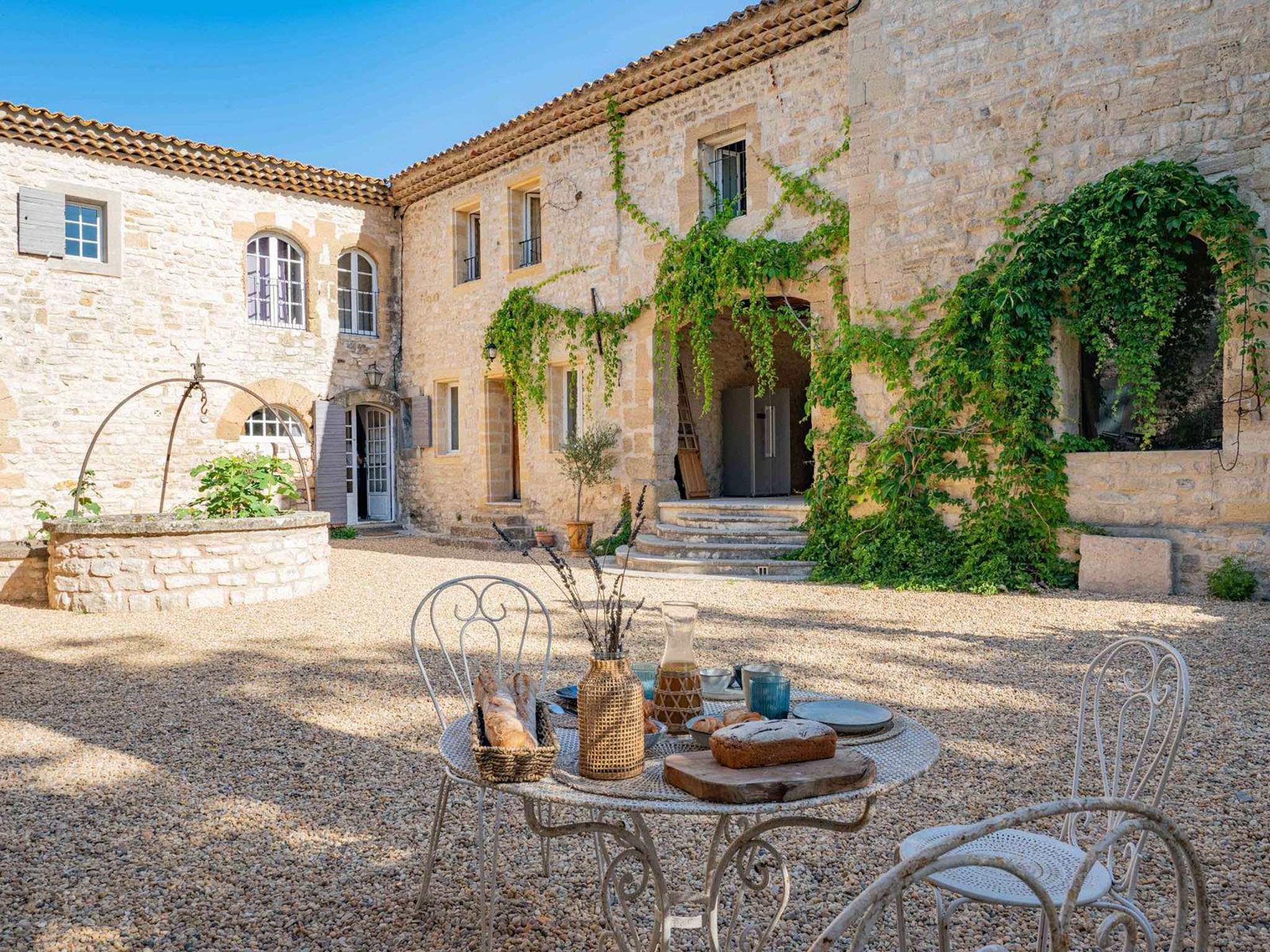 Provencal stone mas courtyard with arched doorways, climbing vines, stone well, and bistro table with French breakfast spread