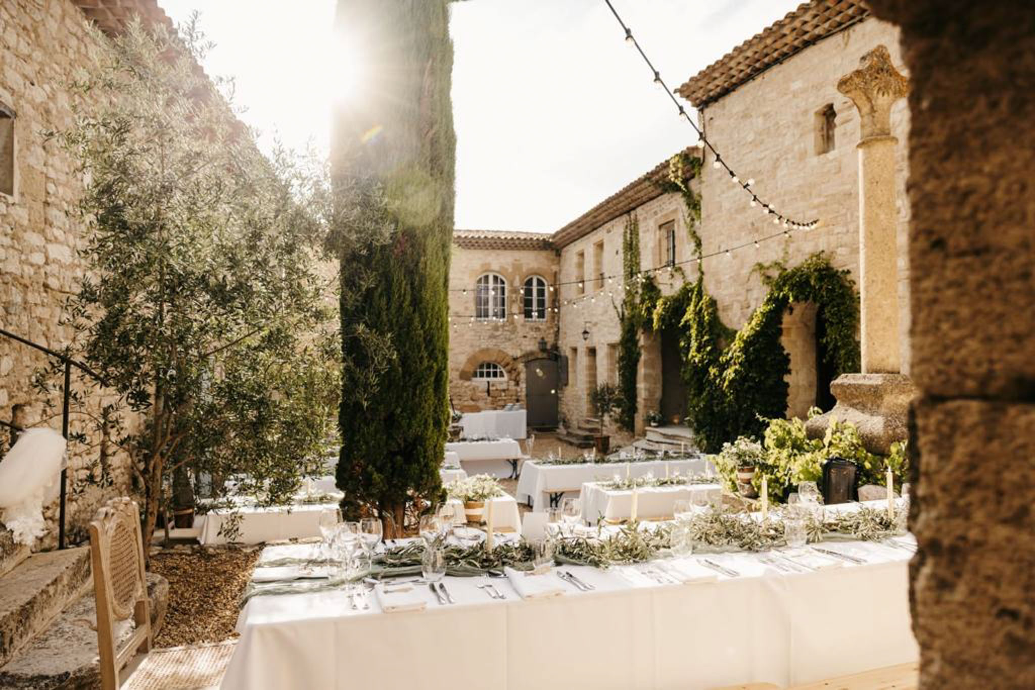 Outdoor reception tables with olive branch runners and taper candles in a stone courtyard with fairy lights