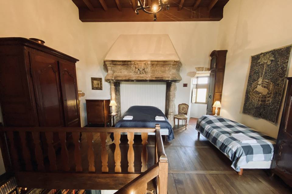 Interior shot of a château bedroom viewed from an elevated mezzanine or staircase landing, showing the room from above at an angle. The room features a large ornate stone fireplace with carved decorative relief as the central focal point, in front of which sits a double bed with a navy blue duvet and white folded towels. A secondary single bed with a blue and grey checked bedspread is positioned to the right. The room is furnished with dark walnut antique armoires, a grandfather clock, a carved wooden chair, and two bedside lamps with warm amber lighting. A large tapestry depicting a unicorn hangs on the right wall, and exposed dark wooden beam ceilings with a wrought-iron chandelier are visible above. The flooring is wide-plank dark hardwood, and the overall style is classic French château with period furnishings. Wide-angle interior shot. Potential venue feature image.