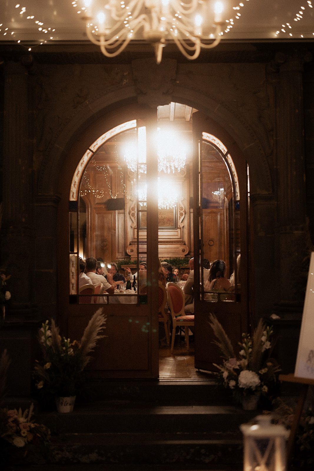 View through arched doors into chateau reception with wood paneling chandelier and white gladioli entry florals